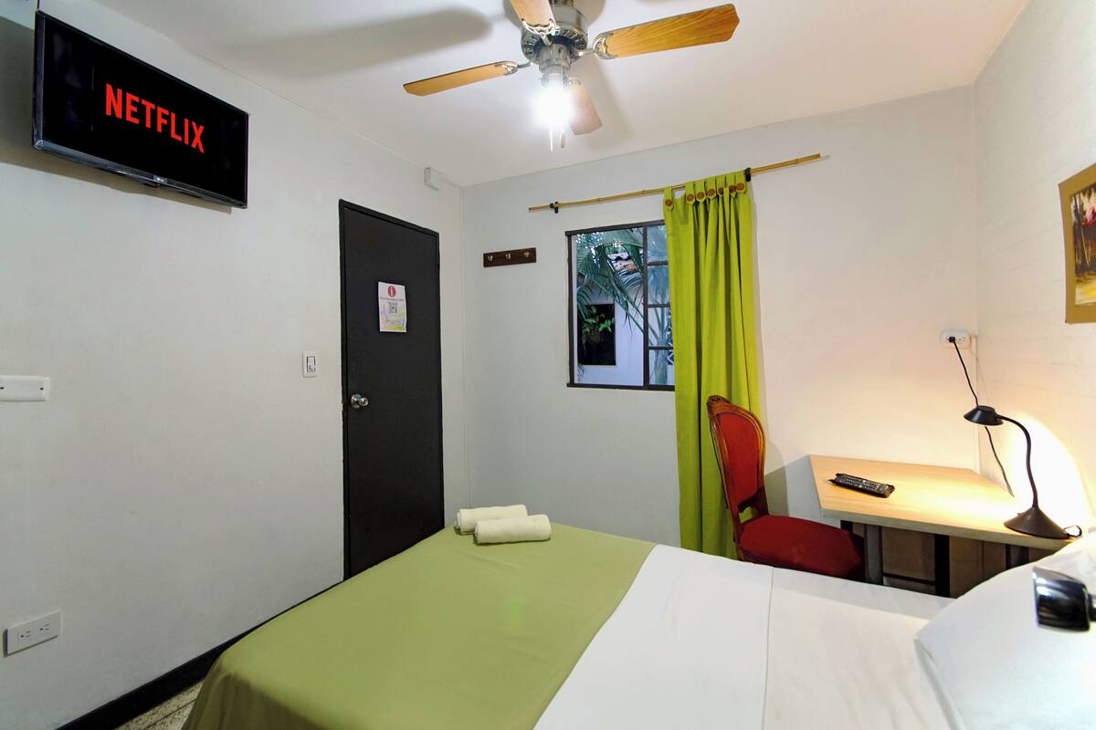 Une chambre d’hôtel équipée d’un lit, d’un bureau, d’une chaise, d’un téléviseur affichant le logo « NETFLIX », d’un ventilateur de plafond et d’une fenêtre avec des rideaux.