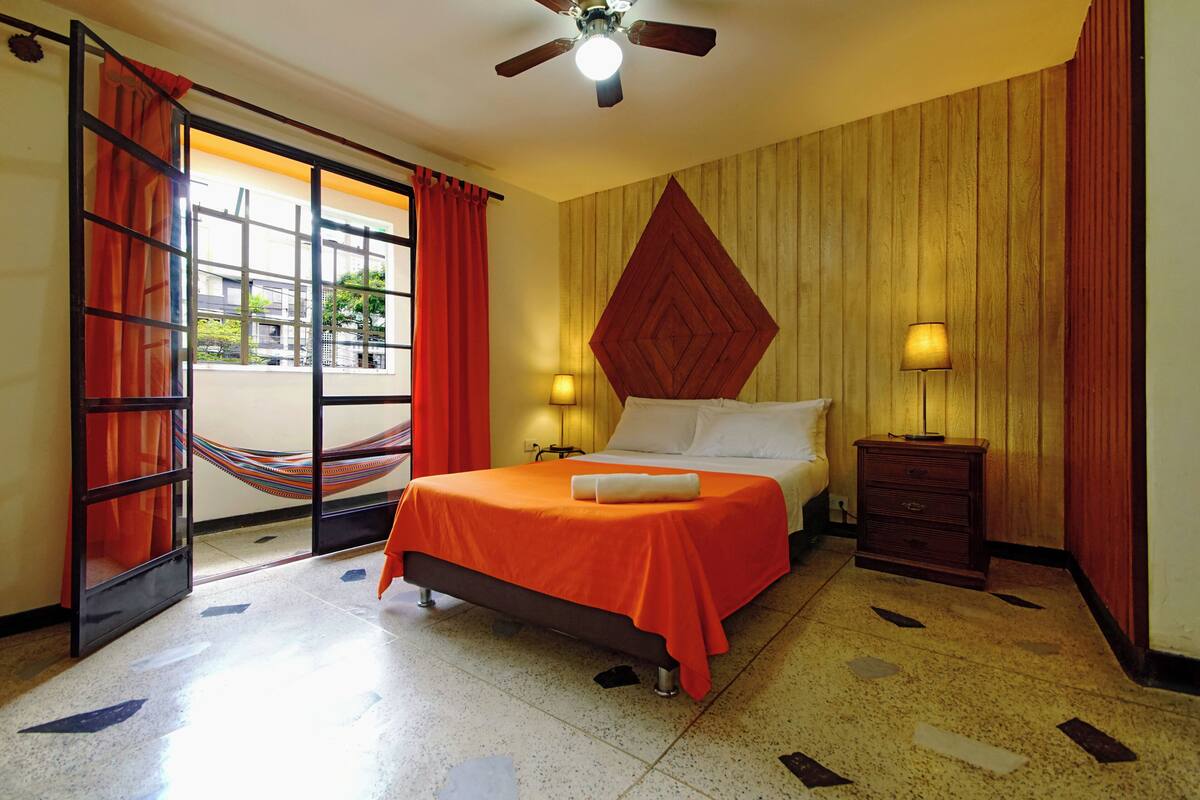 Une chambre d’hôtel avec un lit, une table de chevet, une lampe, un ventilateur de plafond, un hamac et une fenêtre avec des rideaux.