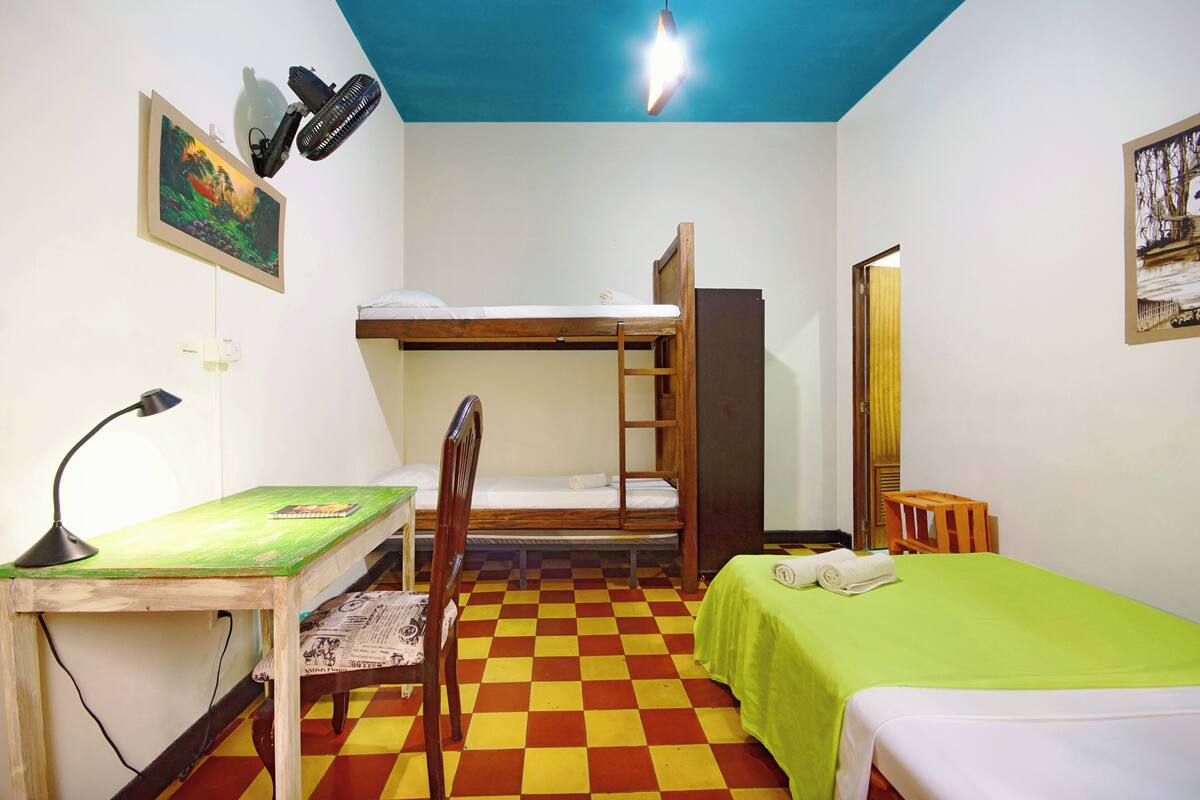 Une chambre de dortoir avec un lit superposé, un bureau avec une chaise, une couverture verte et un sol à carreaux.