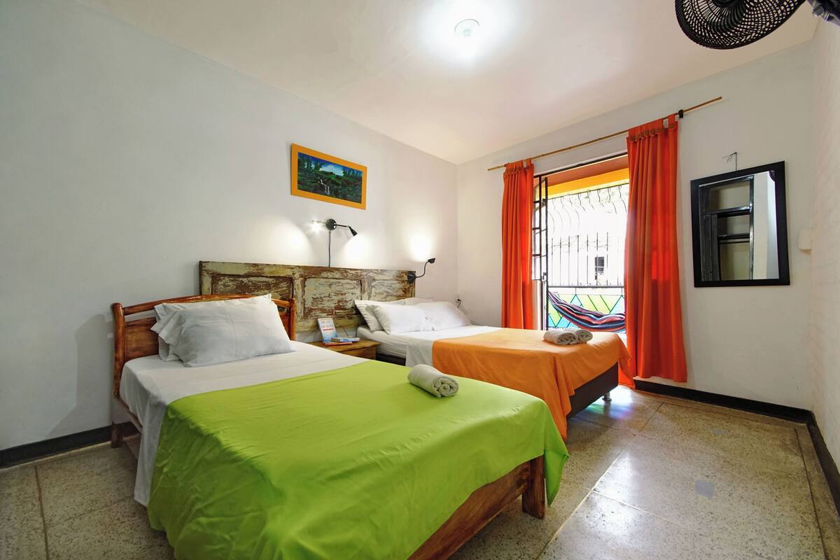 Une chambre avec deux lits, chacun recouvert d’une couverture verte, agrémenté d’oreillers blancs et doté d’une tête de lit en bois. Il y a une fenêtre avec des rideaux orange, un miroir fixé au mur et un tableau au-dessus des lits.
