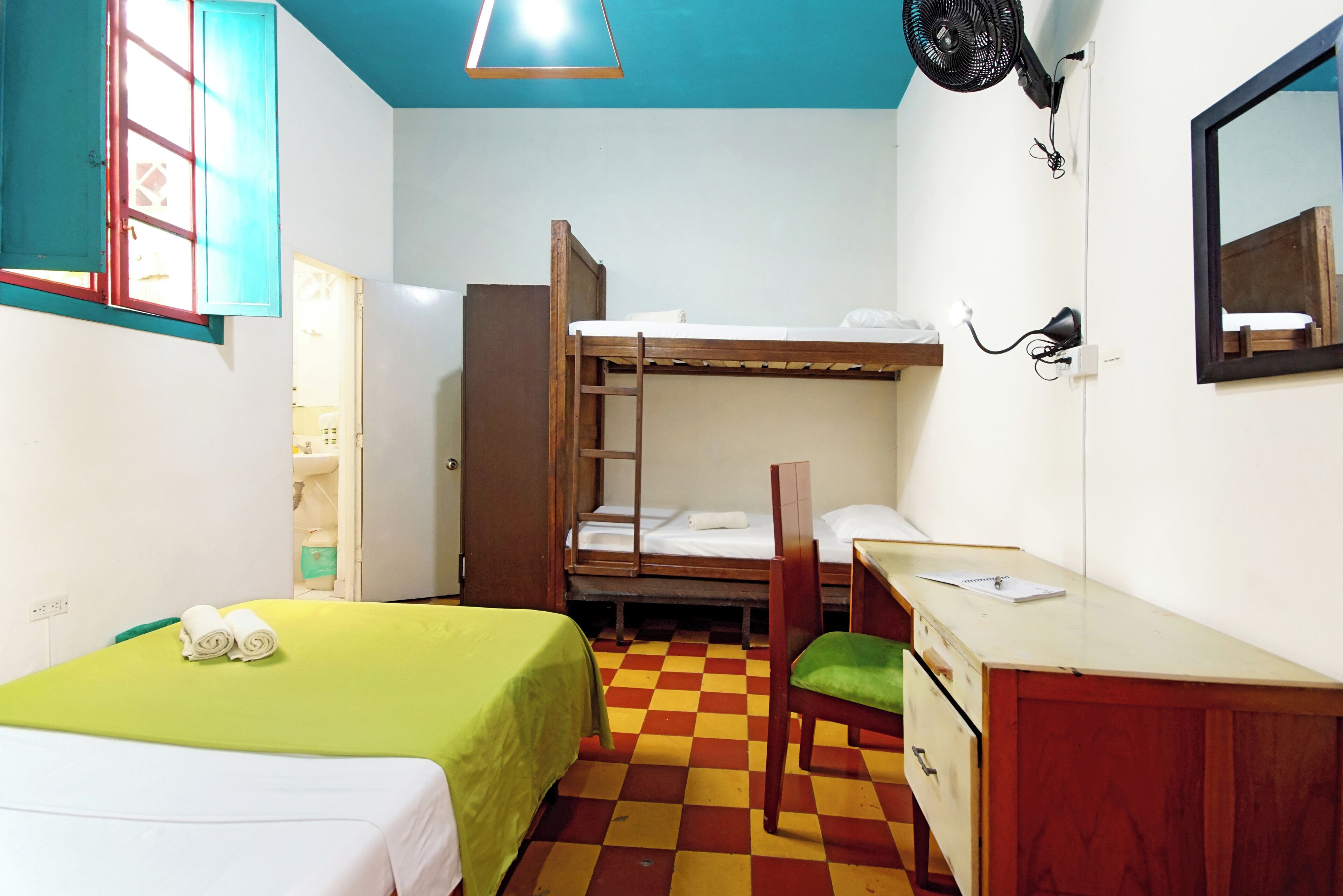 A dormitory room with bunk beds, a desk, a chair, and a fan.