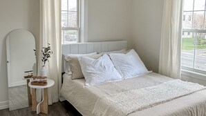 Ein Schlafzimmer mit einem großen Bett, einem Nachttisch mit einer Vase und Kerzen, einer hängenden Rattan-Lampe und Blick ins Freie durch ein Fenster.