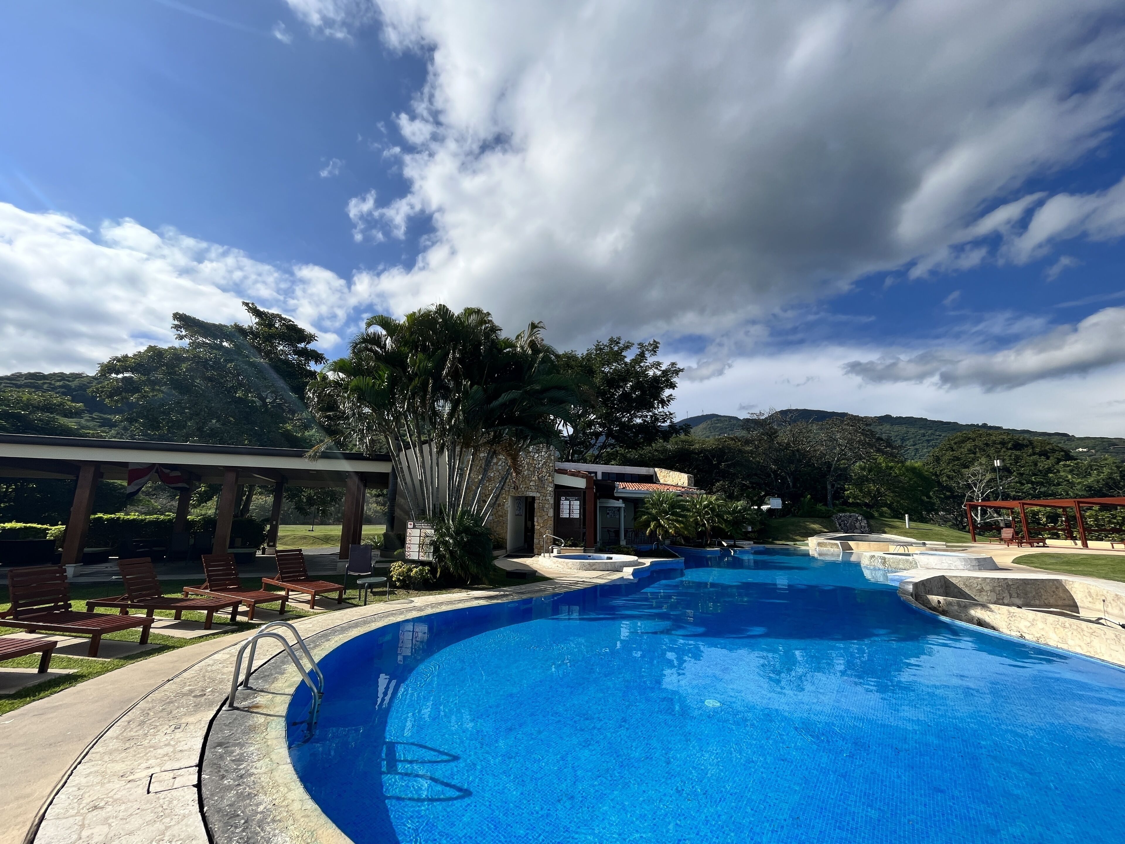 A swimming pool with a clear blue water, surrounded by lounge chairs and a pavilion.