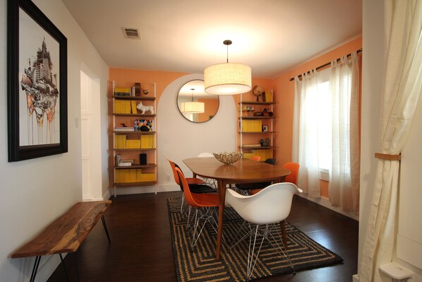 A dining area with a wooden table, white and orange chairs, a woven pendant light, and a mirror on the wall.