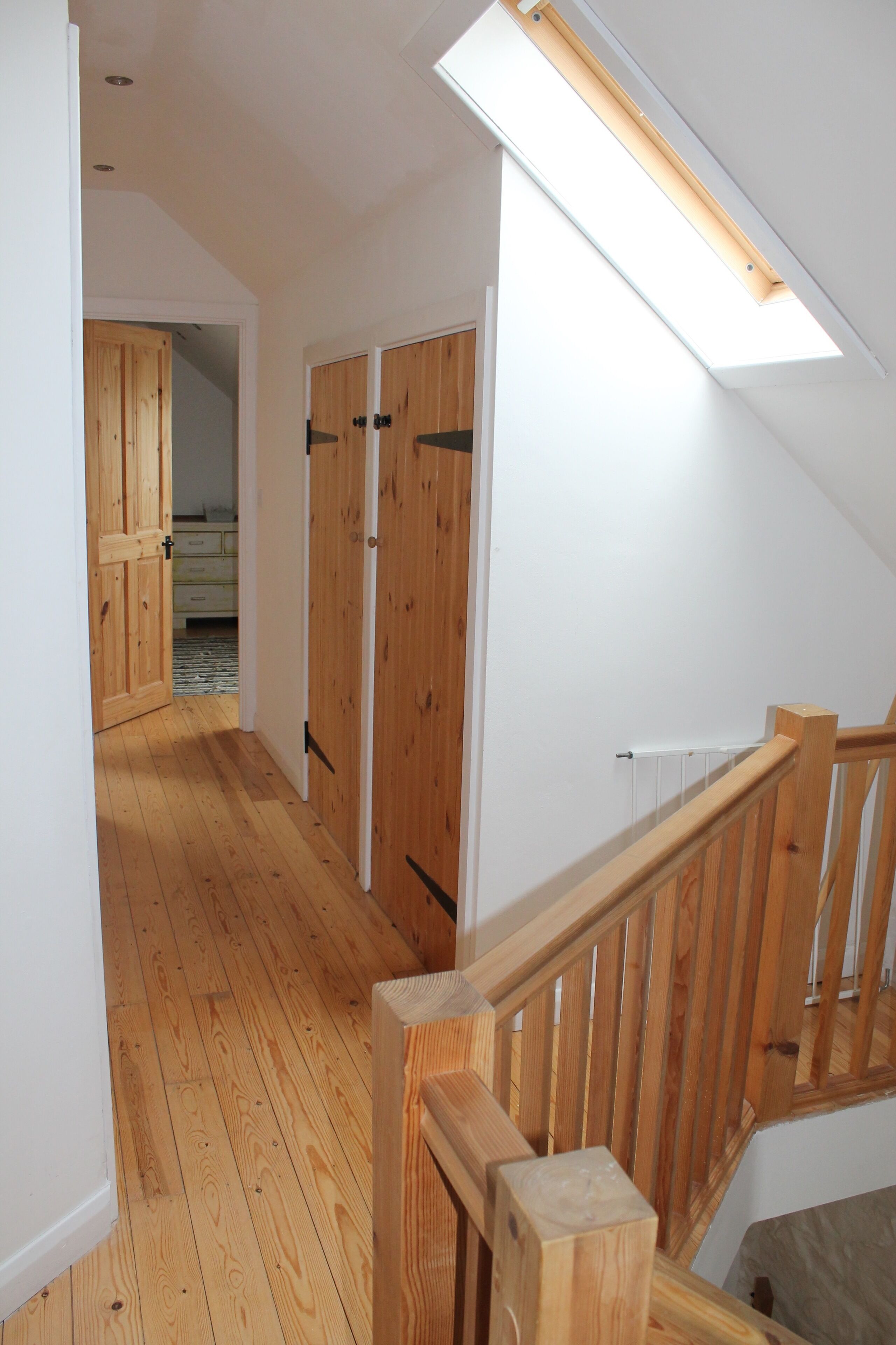 Un escalier en bois avec une rampe, une lucarne et une vue sur une chambre dont les portes sont en bois.