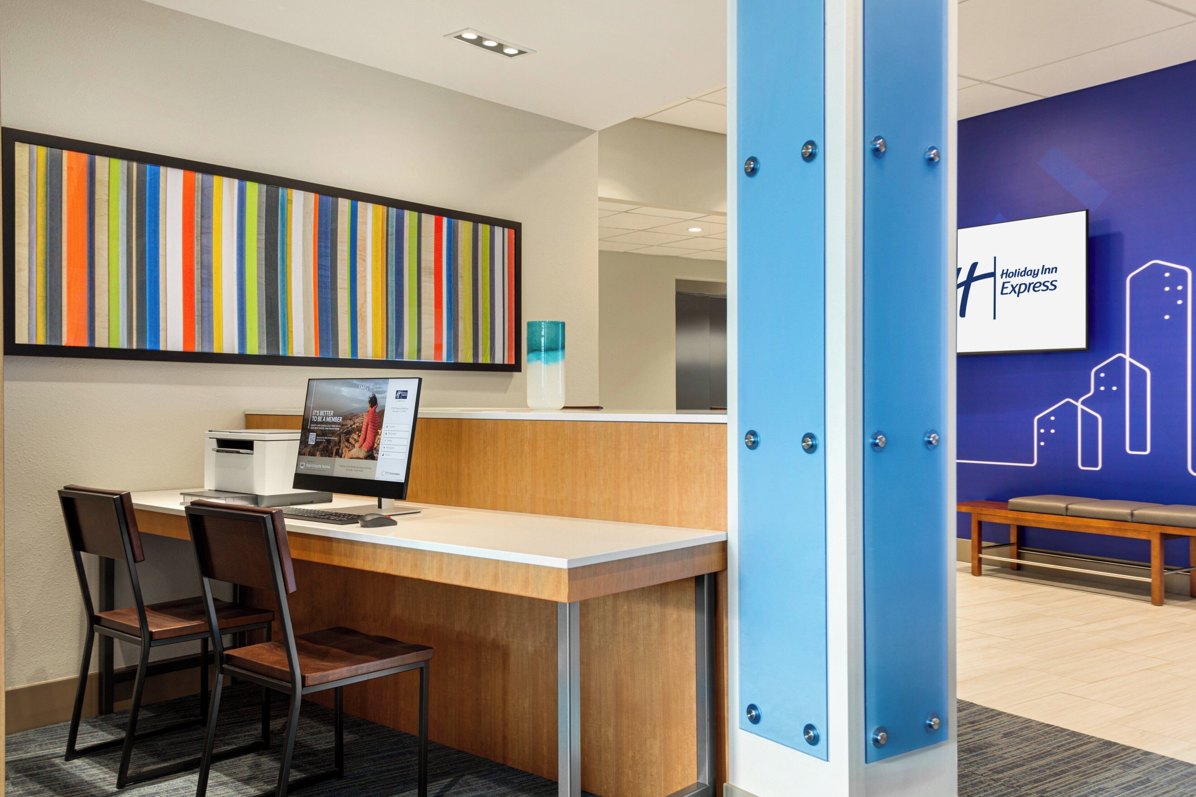 A reception desk with a computer, two chairs, a colorful striped artwork, and a blue wall with a Holiday Inn Express sign.