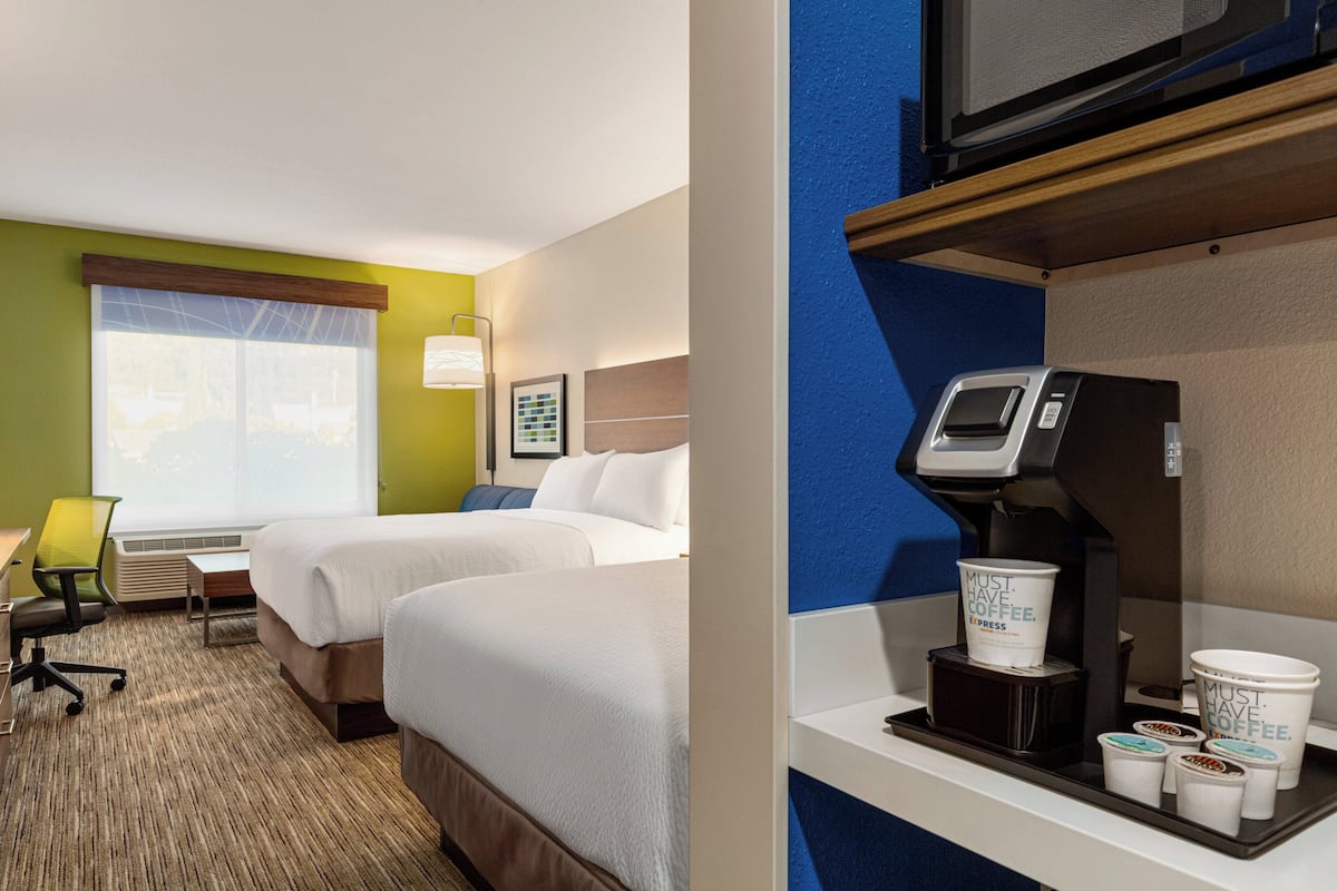 A hotel room with two beds, a desk, a chair, and a coffee machine with disposable cups.