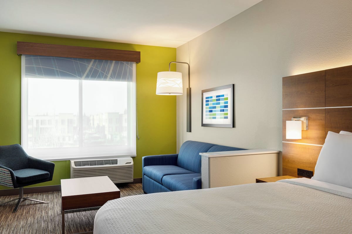 A hotel room with a bed, a desk, a chair, a window with blinds, a wall-mounted lamp, and a framed picture.