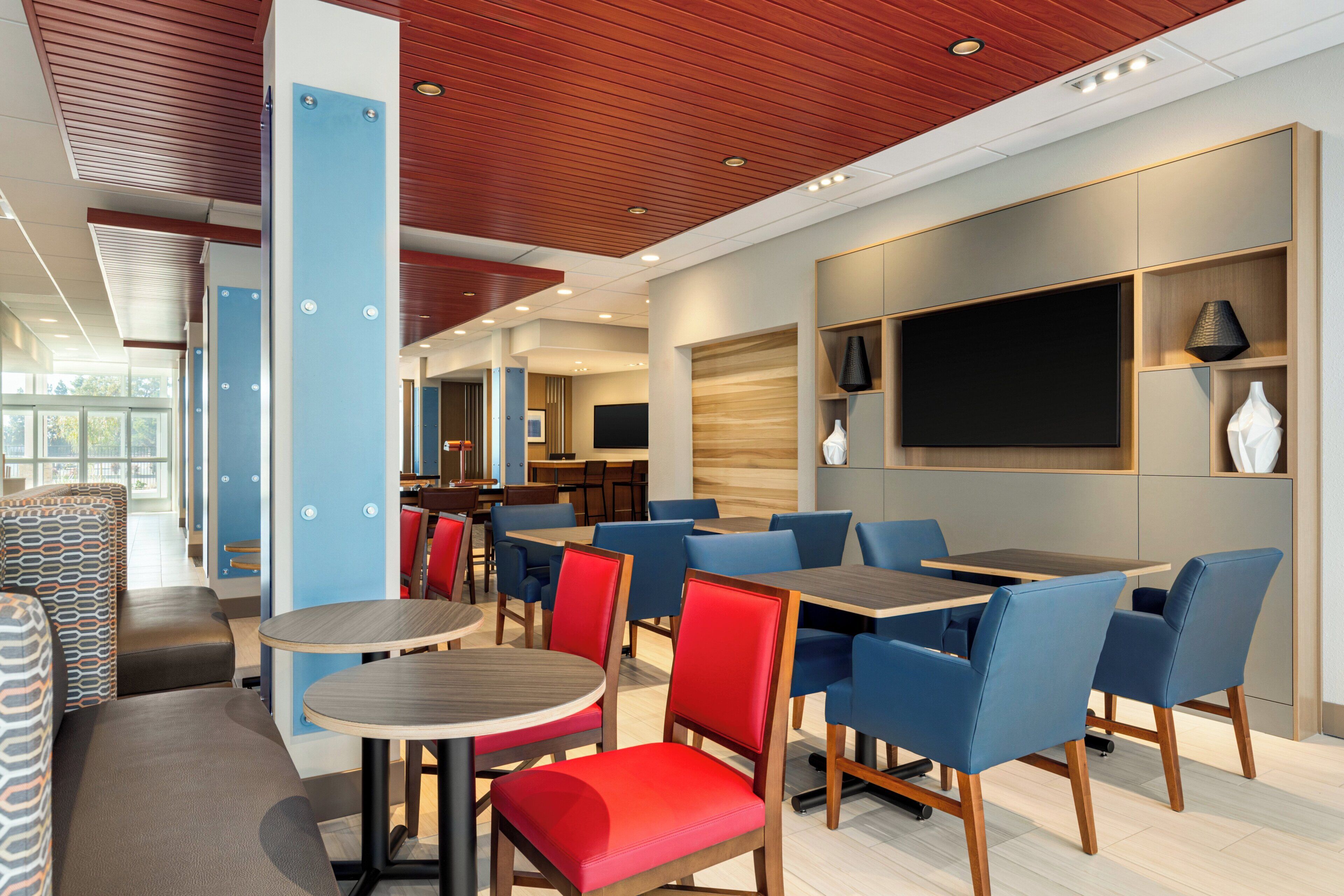 Ein modernes Restaurant mit blauen und roten Sitzgelegenheiten, Holztischen und einem großen Flachbildfernseher an der Wand.