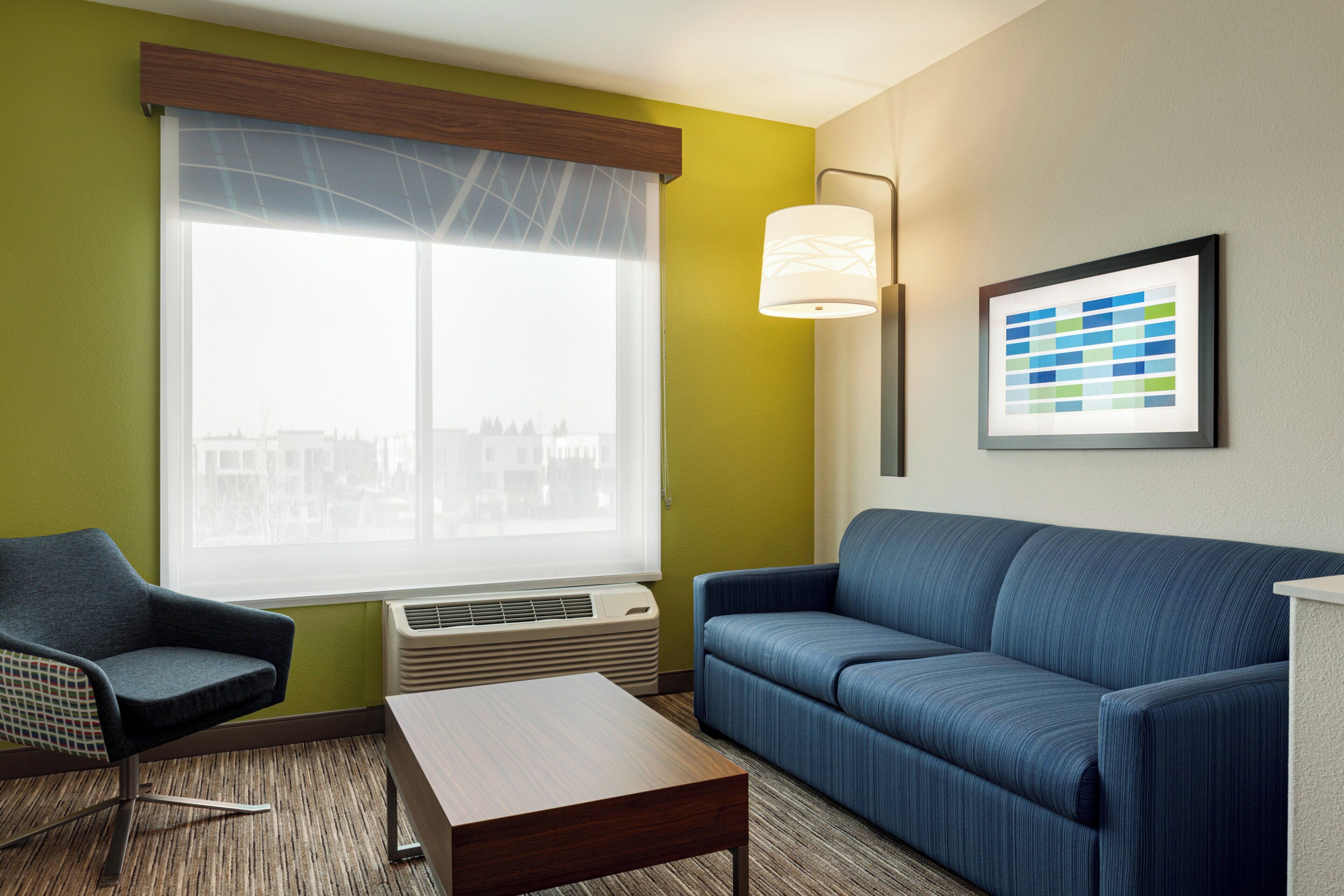 A hotel room with a blue sofa, a dark armchair, a wooden coffee table, a large window with a view of buildings, and a framed picture on the wall.