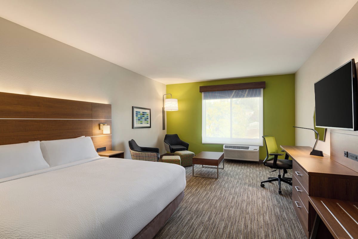 A hotel room with a large bed, a desk, a chair, a small table, a lamp, a TV, and a window with blinds.
