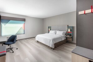 A modern hotel room with a large bed, a desk, and a chair. There is a window with blinds, a nightstand with a lamp, and a wall-mounted shelf. - avid hotel North Platte by IHG (North Platte)