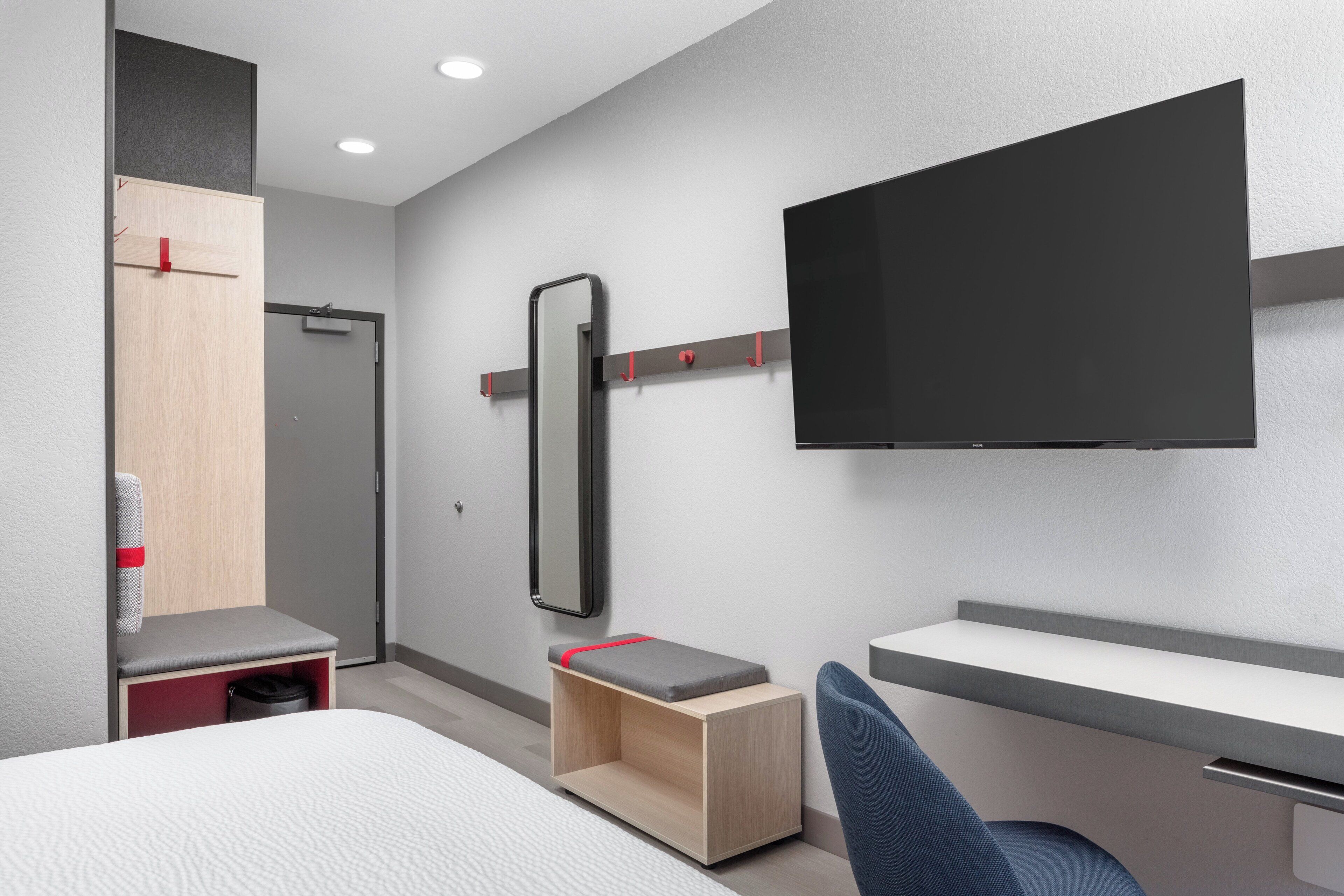 Une chambre d’hôtel moderne équipée d’un lit, d’un bureau, d’une chaise, d’une télévision, d’une armoire et d’un miroir.