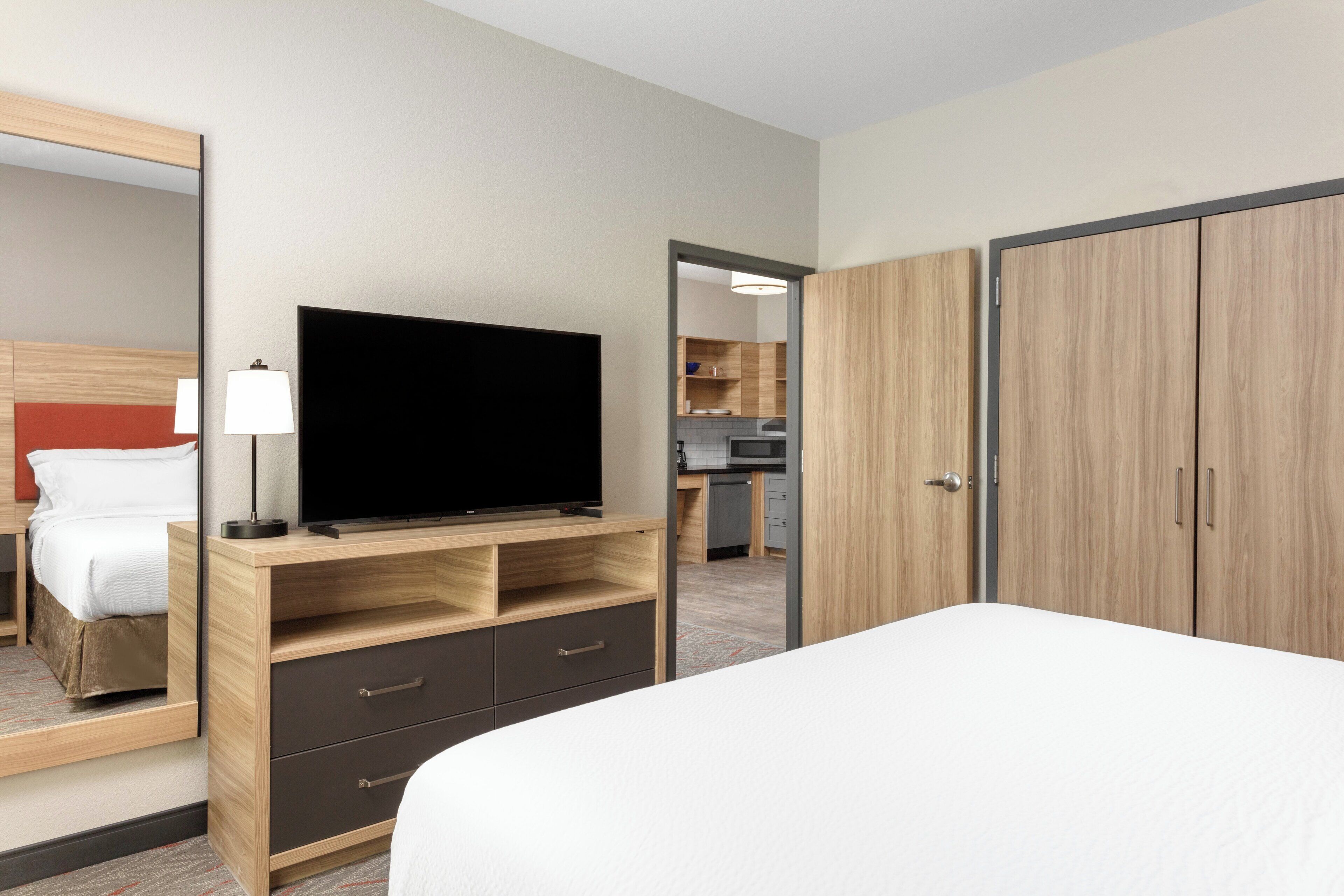 Une chambre d’hôtel avec un lit, un téléviseur posé sur un meuble en bois, un miroir, une armoire et une vue sur la cuisine.