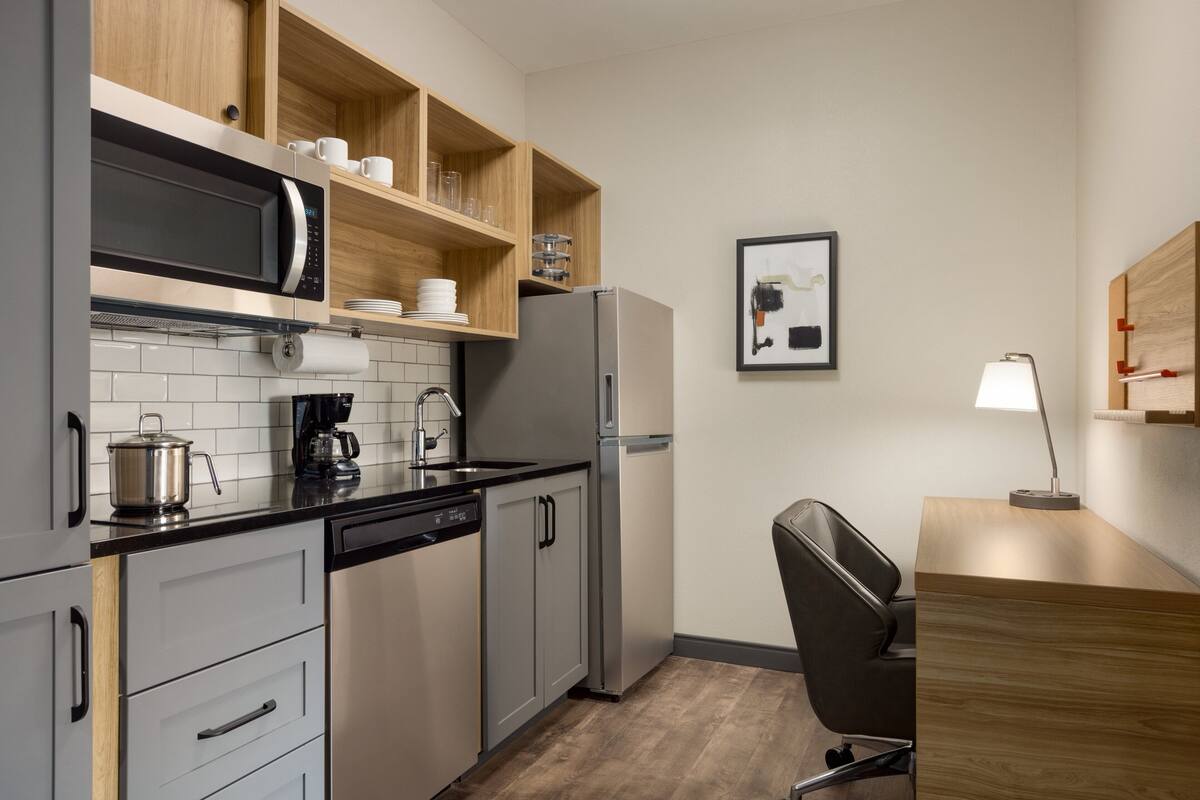 A modern kitchen with a microwave, coffee maker, and refrigerator. There is a wooden shelf above the sink with cups and glasses. A framed picture hangs on the wall, and a desk with a lamp is in the corner.