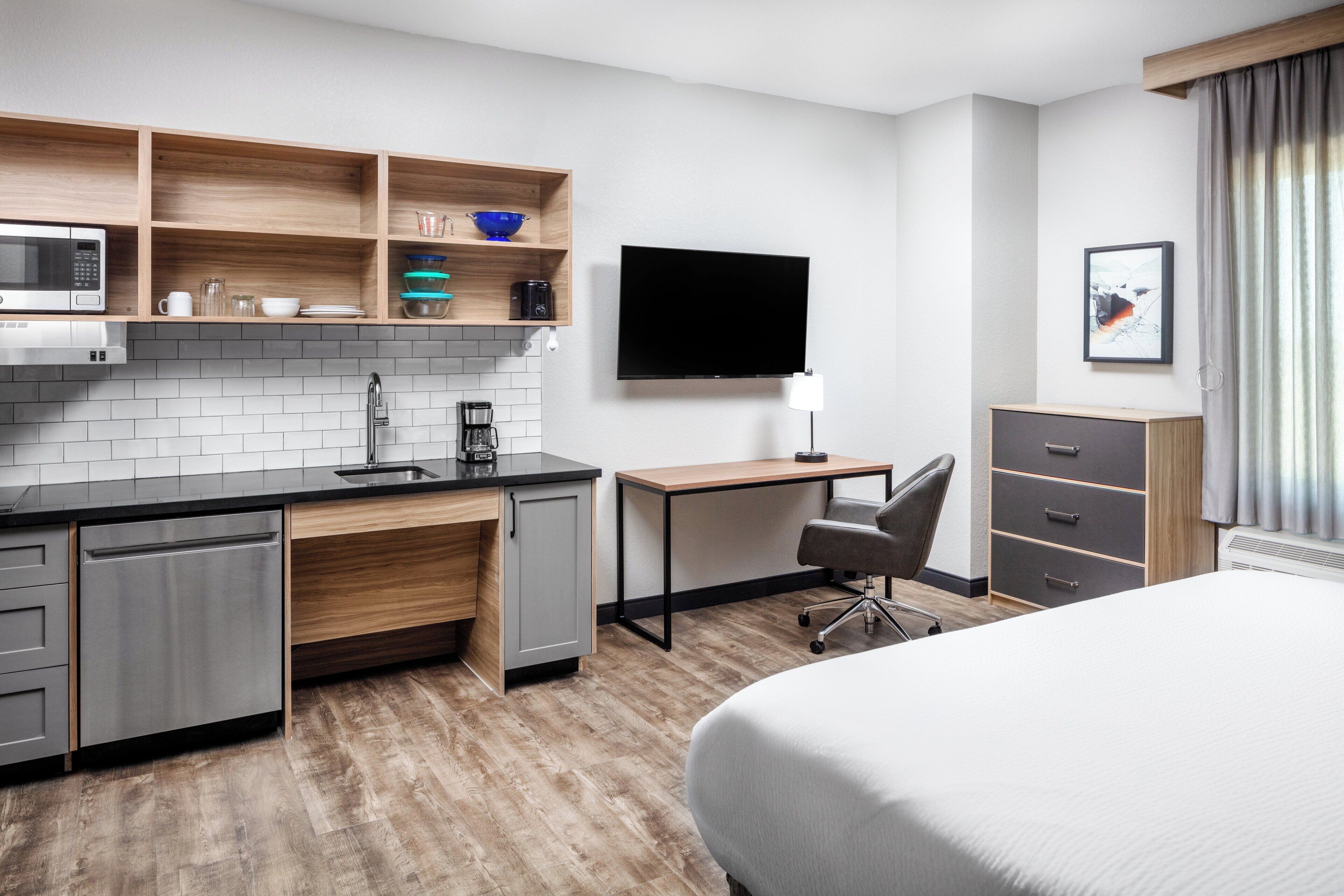A modern hotel room with a kitchenette, a flat-screen TV, a desk, and a bed.