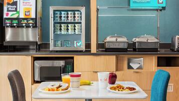 Ein Frühstücksbereich mit Automaten, Kühlschrank und Mikrowelle. Zwei Stühle und ein Tisch mit Frühstücksangeboten.