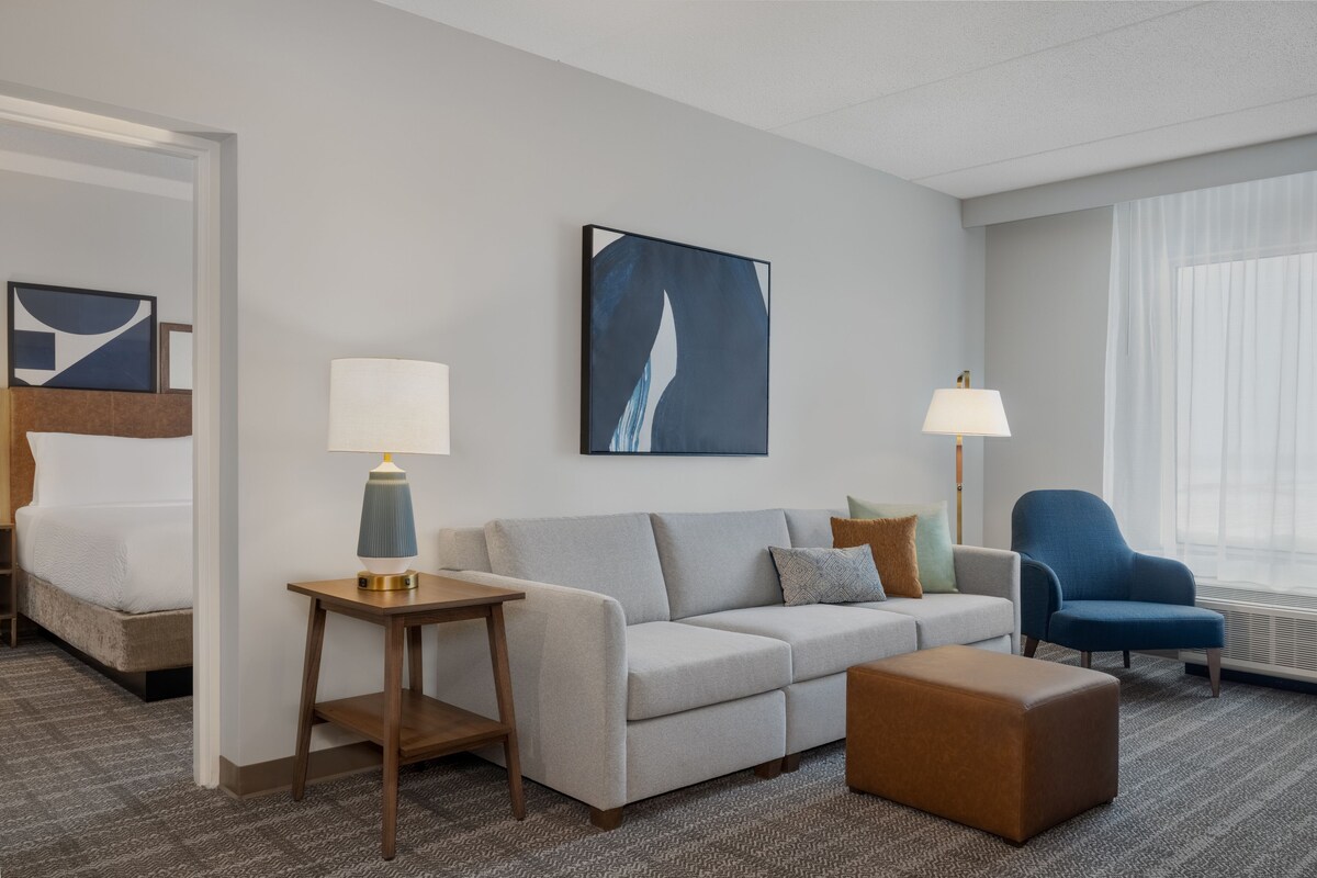 A hotel room with a bed, a gray sofa, a blue armchair, a small table with a lamp, and a large abstract painting on the wall.