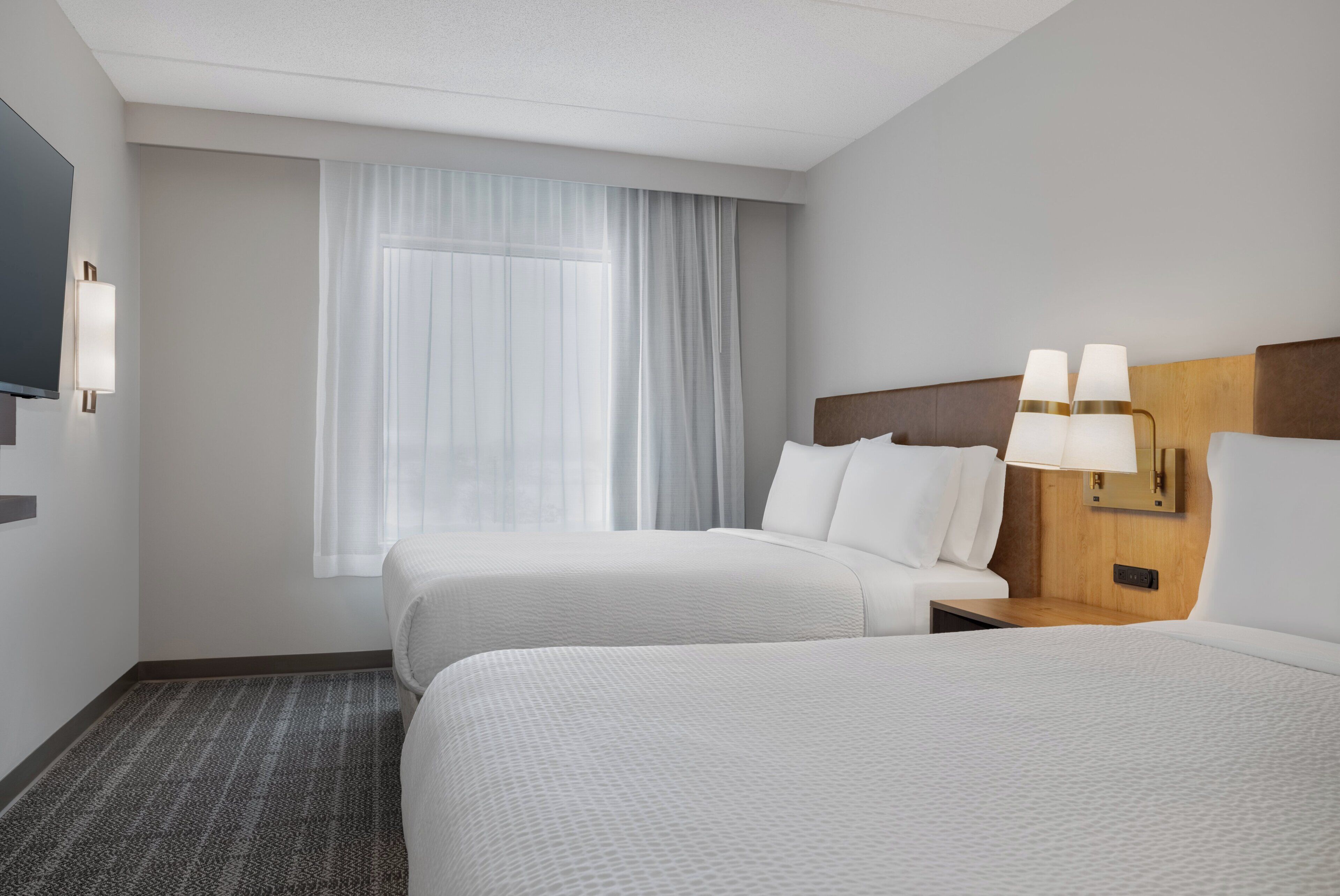 A hotel room with two beds, a large window with sheer curtains, a flat-screen TV mounted on the wall, and a bedside table with a lamp.