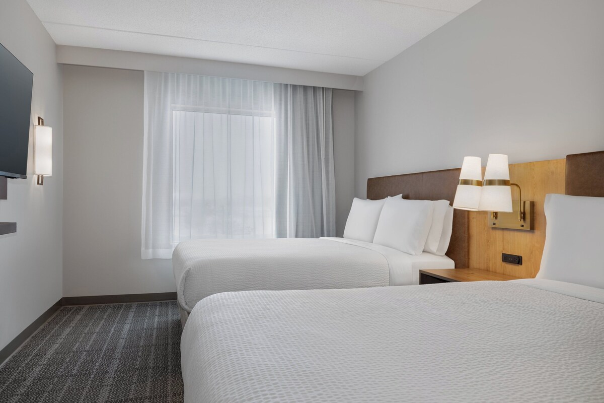A hotel room with two beds, a large window with sheer curtains, a flat-screen TV, and a bedside table with lamps.