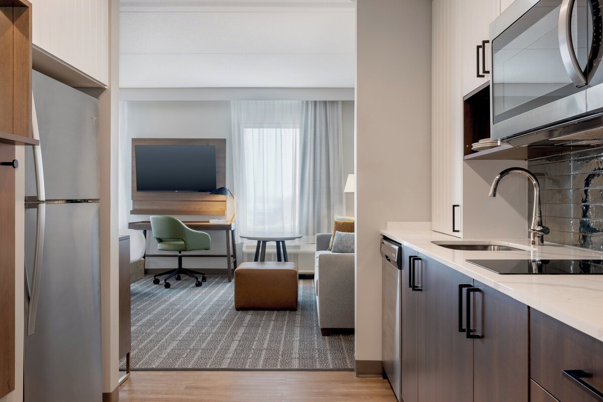 A modern hotel room with a kitchenette, a dining area, and a seating area with a TV.