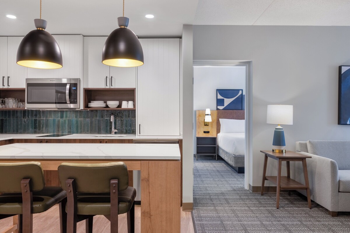 A modern hotel room with a kitchen area, a dining table with chairs, a sofa, a lamp, and a bed.