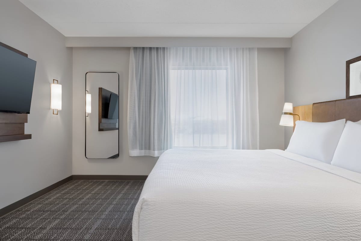 A hotel room with a large bed, a flat-screen TV mounted on the wall, a mirror, a window with sheer curtains, and a bedside lamp.