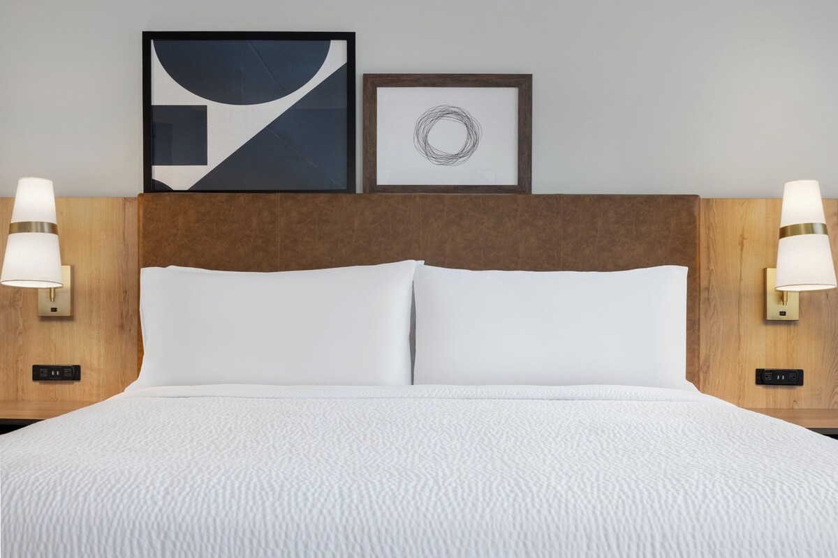 A hotel room with a large bed, two wall-mounted lamps, and two framed artworks on the wall.