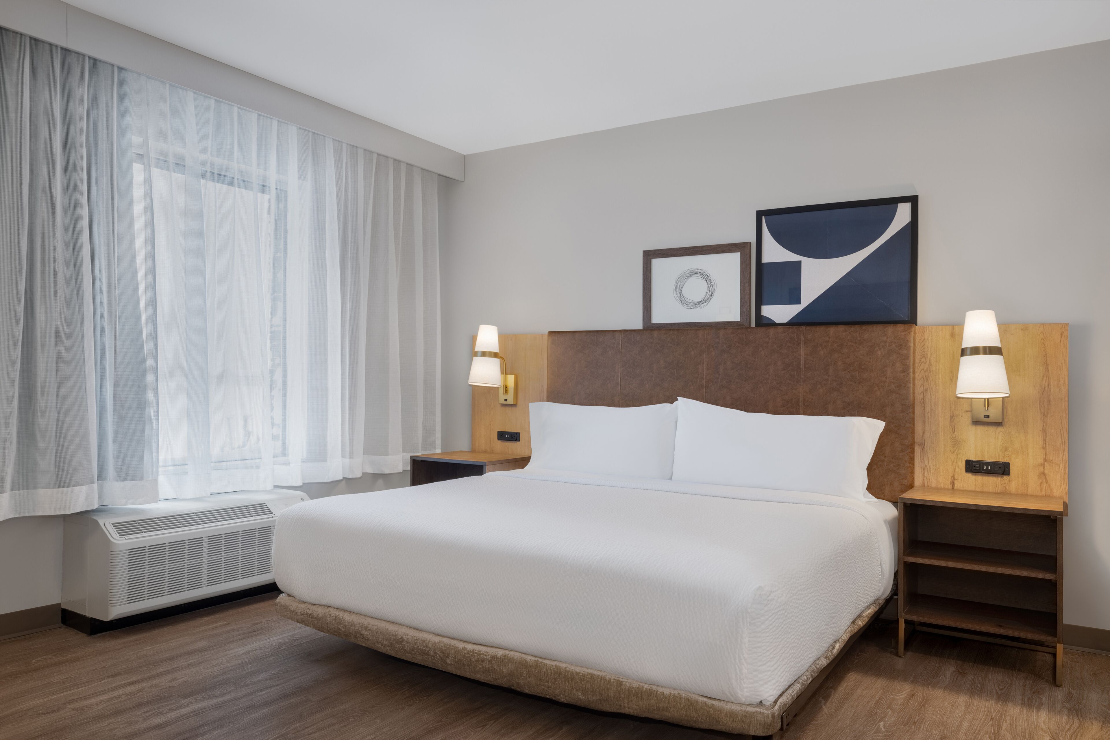 A hotel room with a large bed, two bedside tables with lamps, a window with sheer curtains, and a wall-mounted artwork.