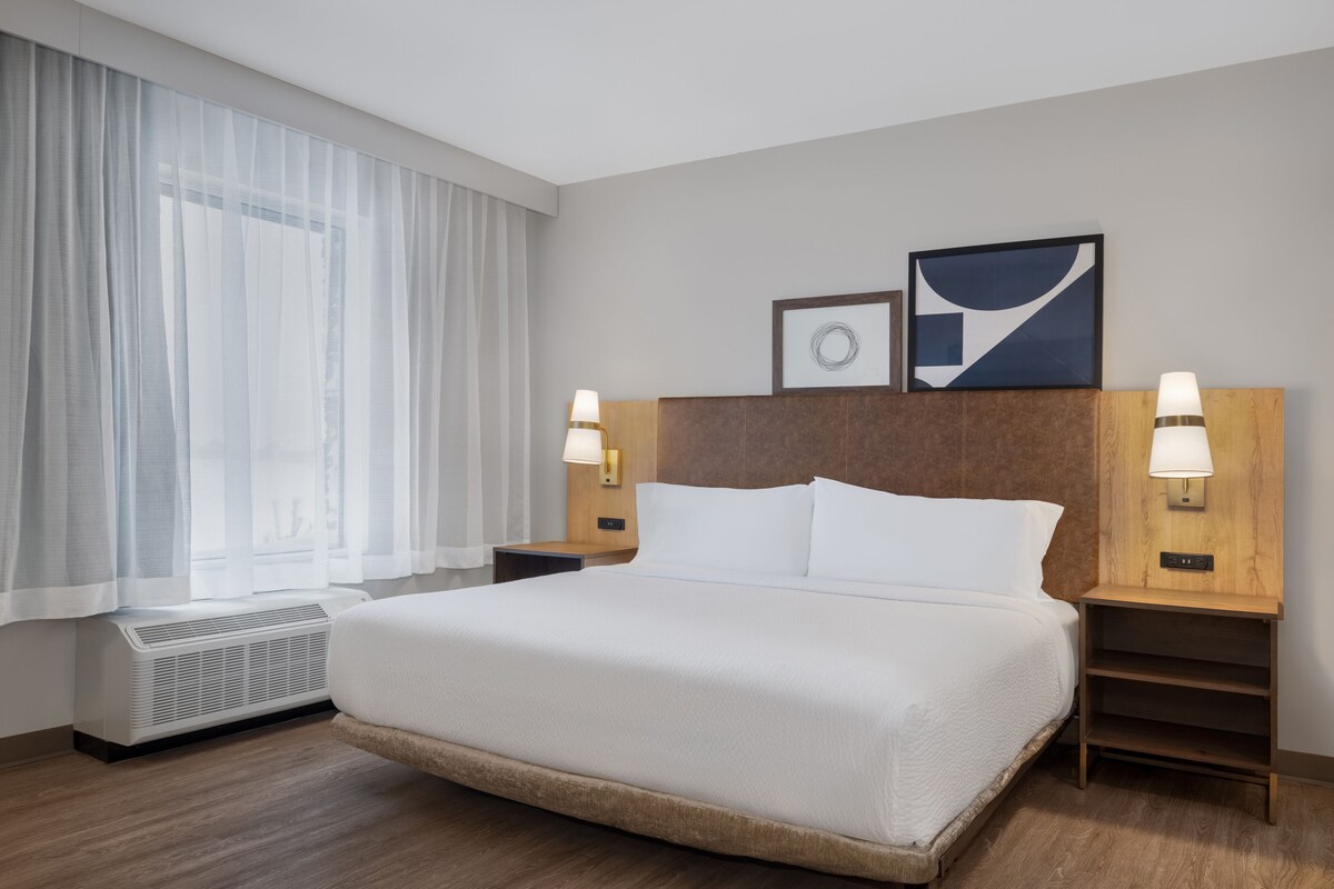 A hotel room with a large bed, two bedside tables with lamps, a window with sheer curtains, and a wall-mounted artwork.