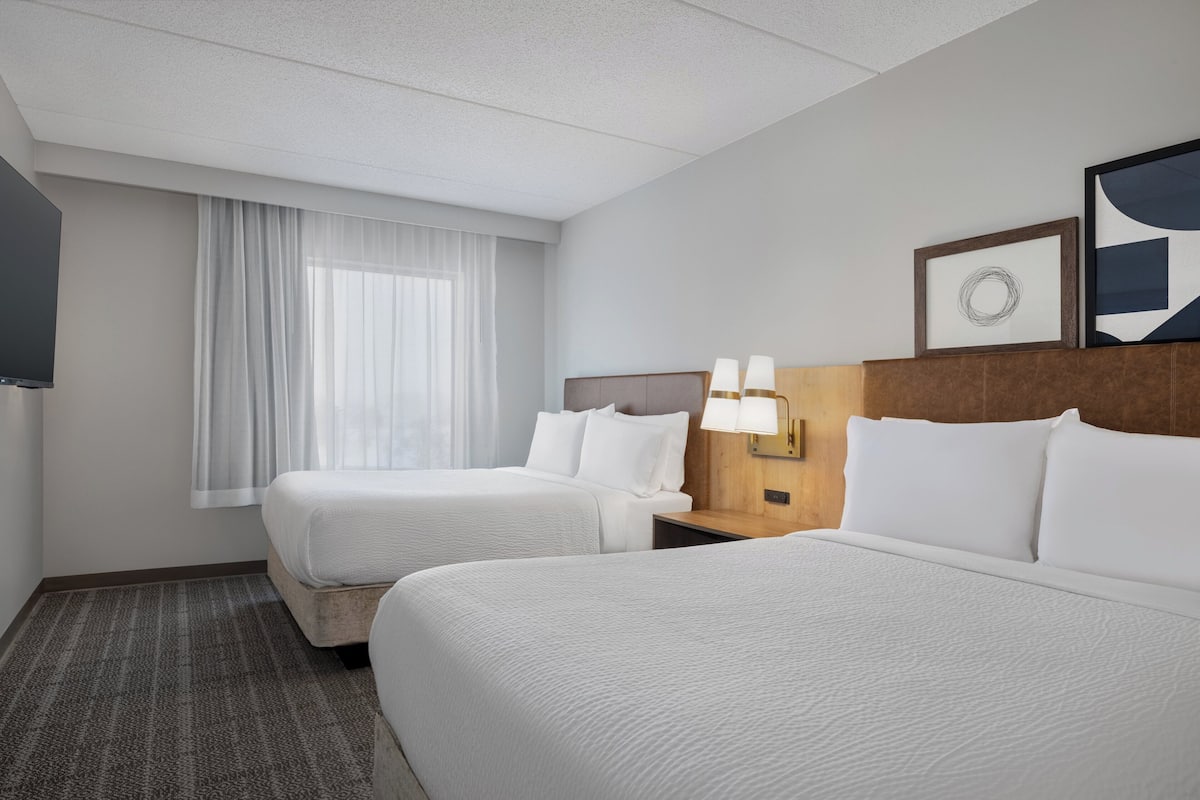 A hotel room with two beds, a TV, a window with curtains, a wall-mounted lamp, and framed artwork.