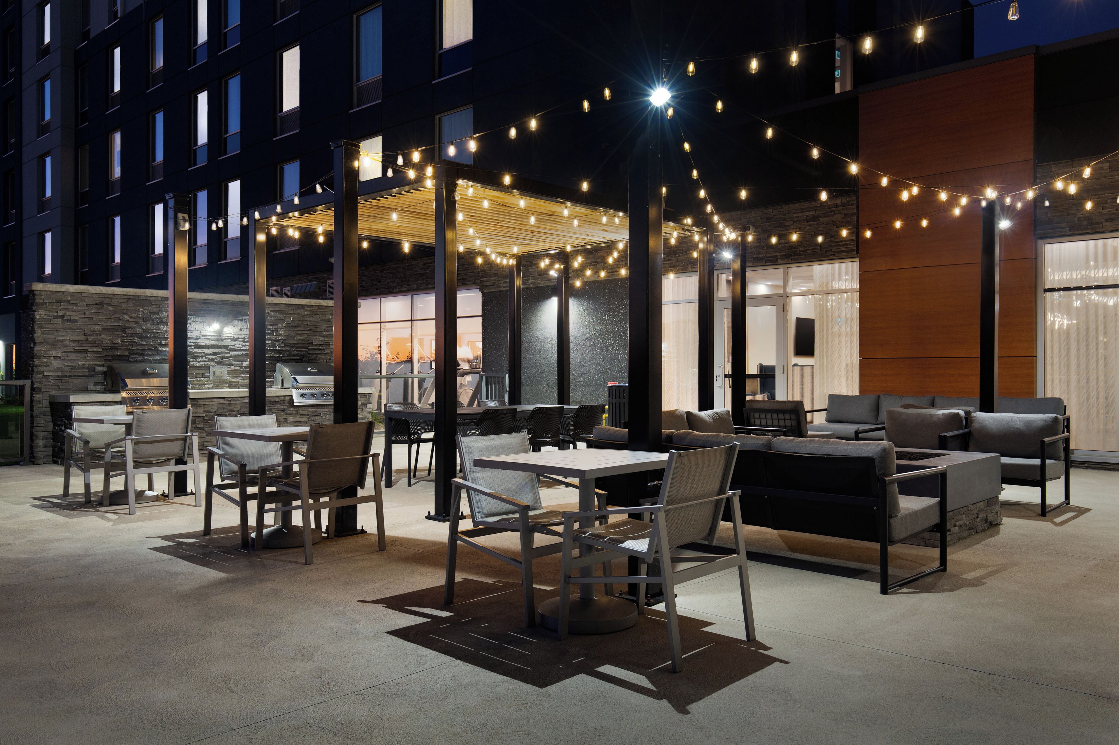 An outdoor patio area with tables and chairs, string lights, and a view of a building in the background.