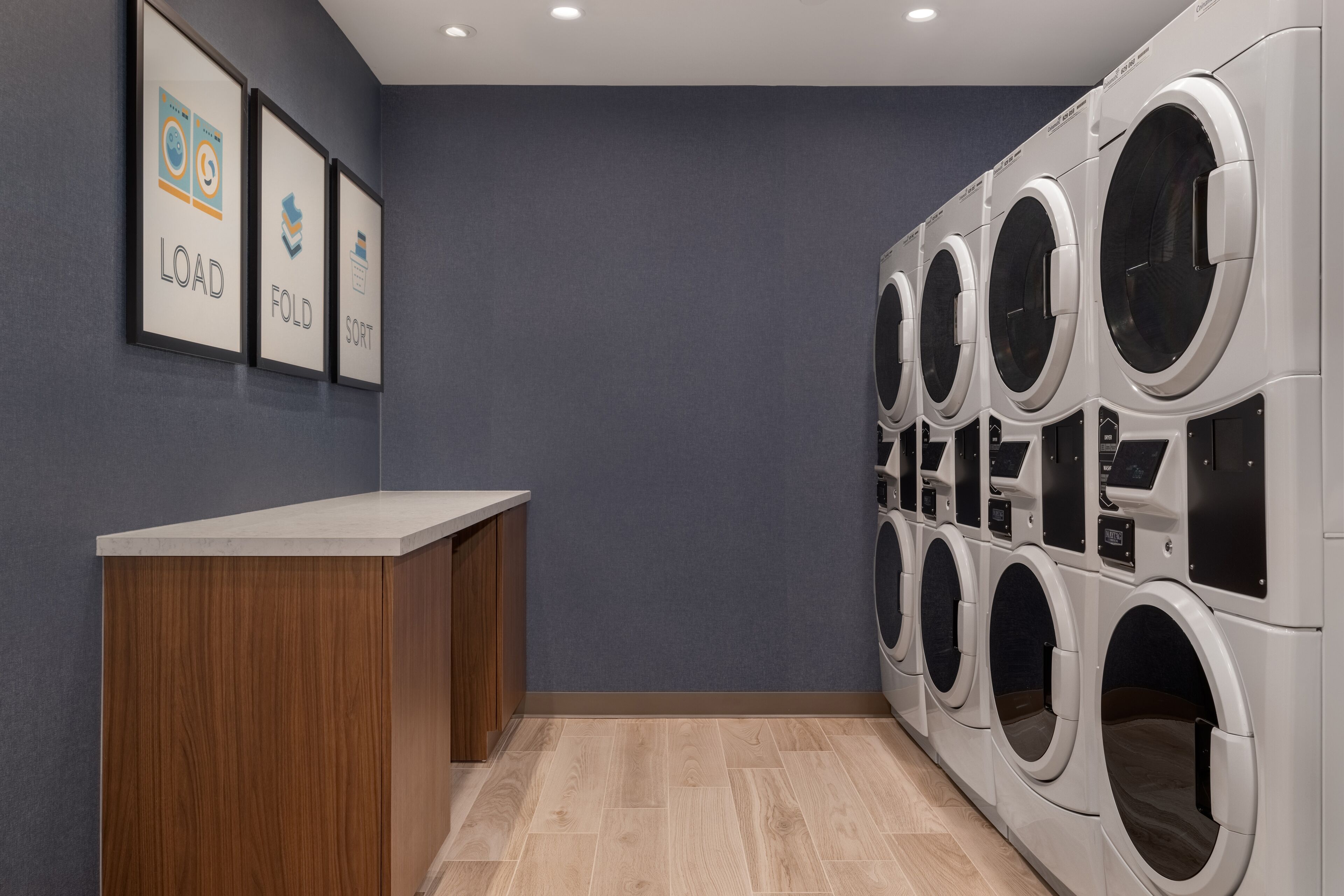 Une buanderie équipée de plusieurs machines à laver, d’un comptoir en bois et de panneaux fixés au mur indiquant les fonctions « CHARGEMENT », « PLIAGE » et « TRI ».