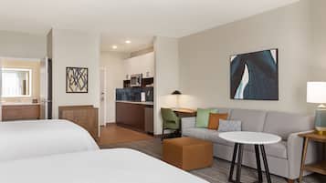A hotel room with two beds, a sofa, a small table, a lamp, a kitchen area, and artwork on the walls.