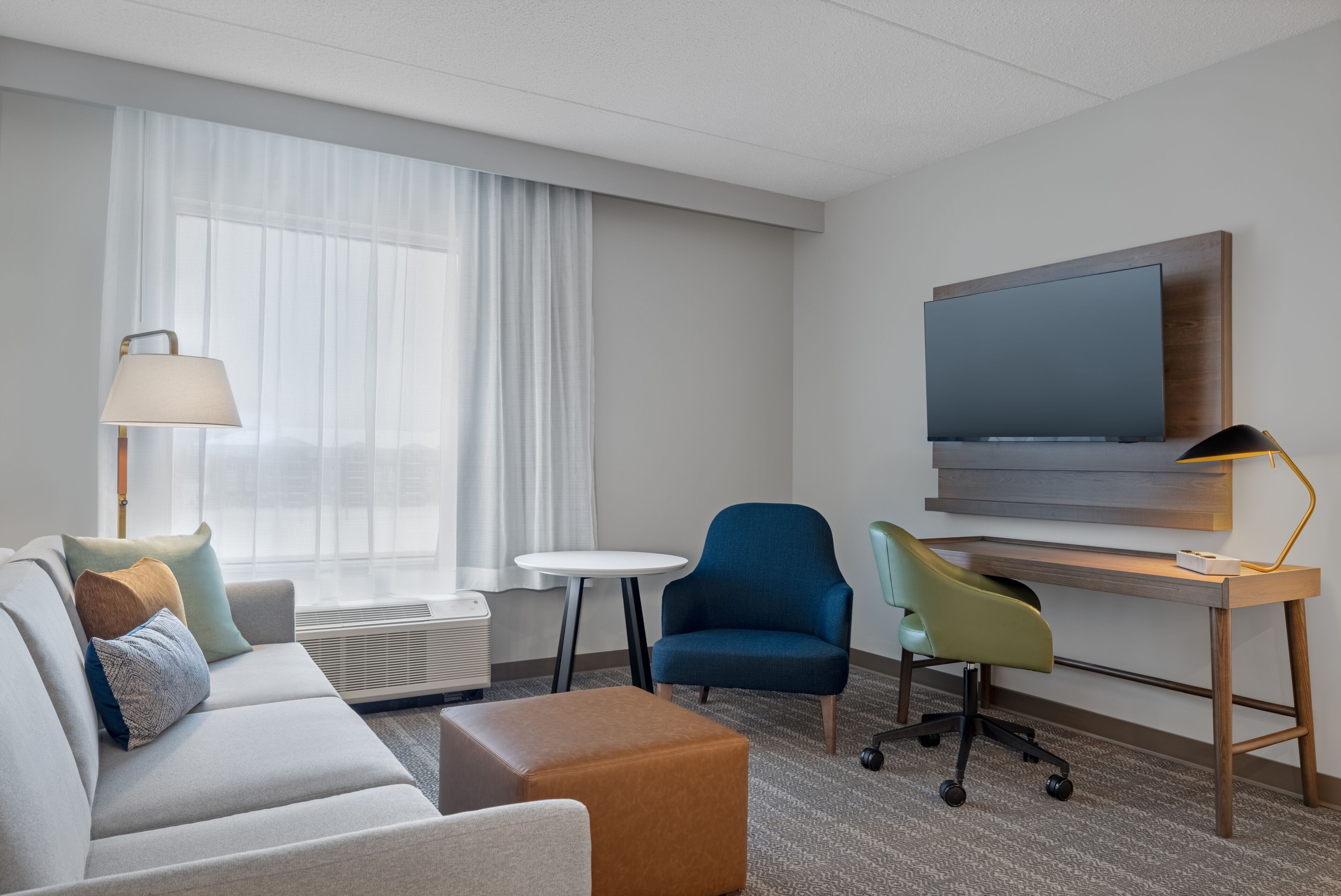 A modern hotel room with a sofa, a small round table, a blue armchair, a green chair, a wooden desk with a flat-screen TV, and a lamp.