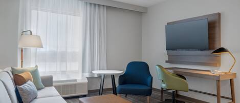 A modern hotel room with a sofa, a small round table, a blue armchair, a green chair, a wooden desk with a flat-screen TV, and a lamp.