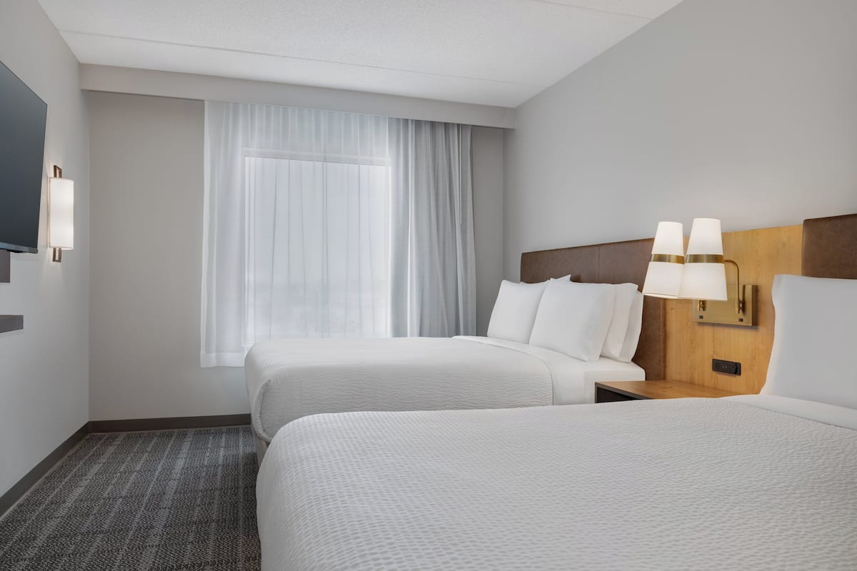 A hotel room with two beds, a large window with sheer curtains, a flat-screen TV, and a bedside table with lamps.