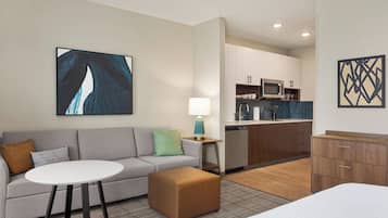 A modern hotel room with a grey sofa, a round white table, a wooden cabinet, and a kitchen area with a microwave and dishwasher.