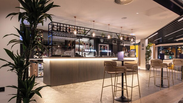 A modern bar area with a herringbone wood floor, bar stools, and a variety of bottles and glasses on shelves.
