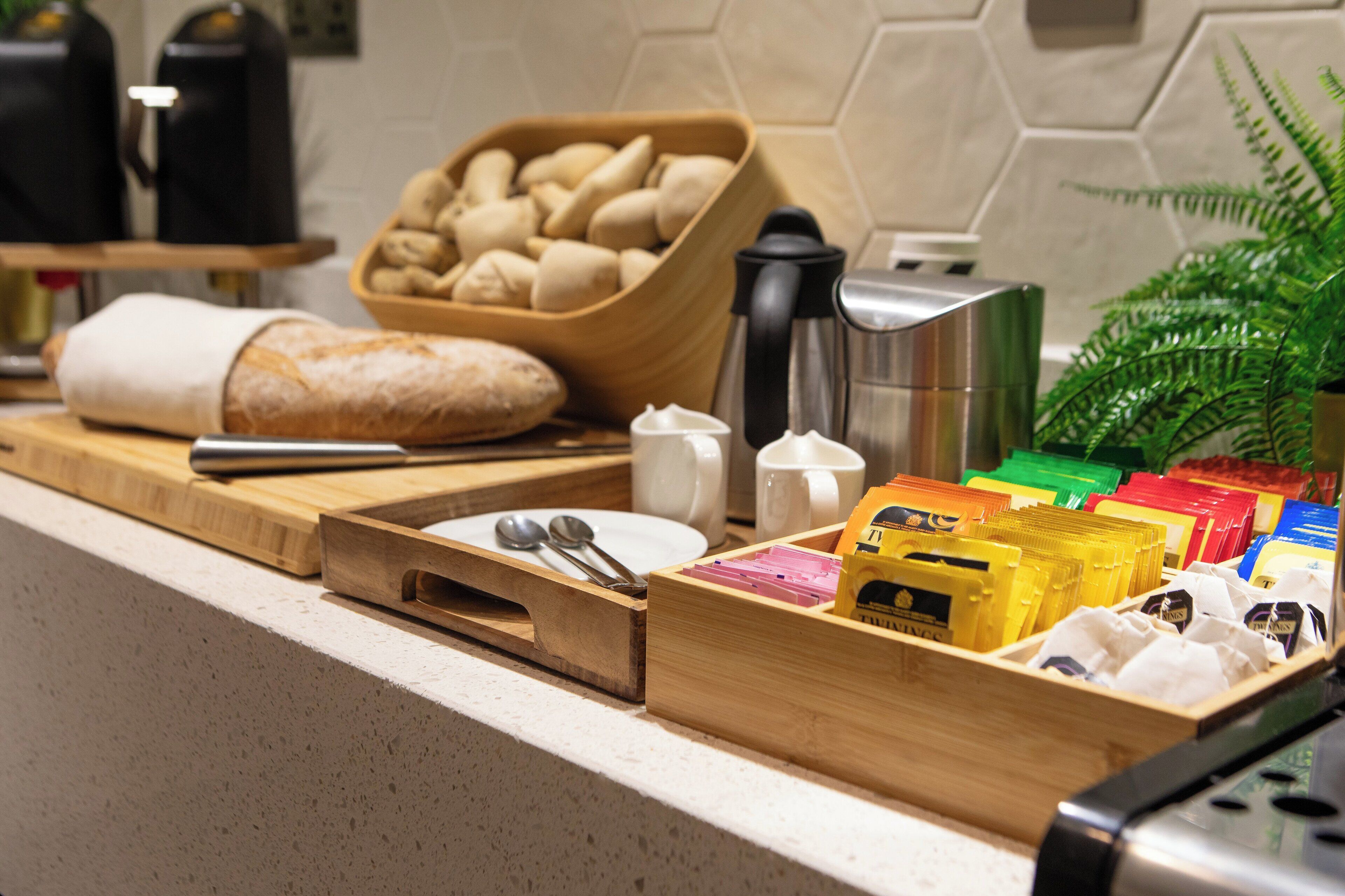 Una encimera de cocina con una hogaza de pan, un bol de panecillos, una cafetera y un organizador de cajones con varios objetos.