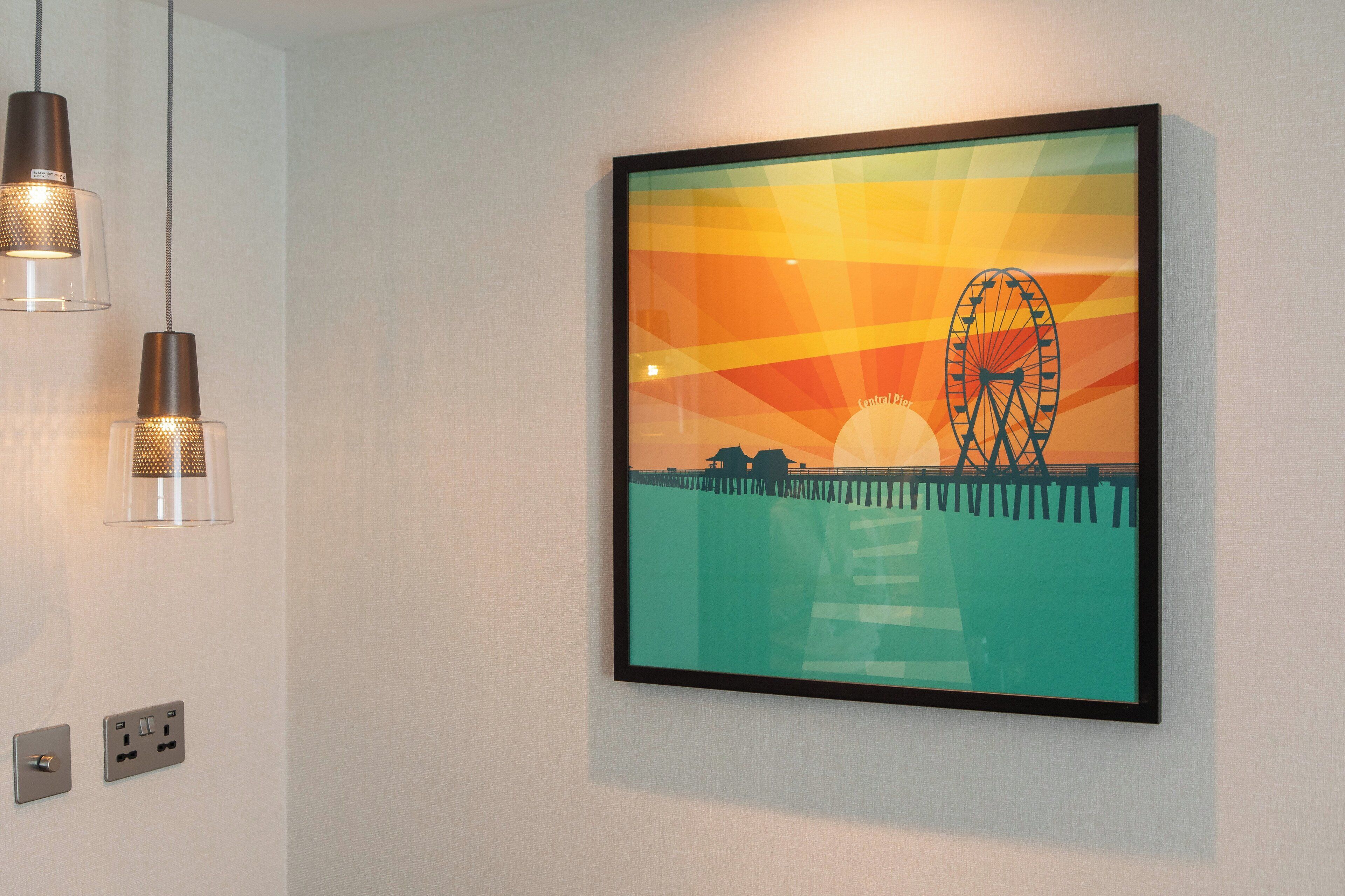 Una obra de arte enmarco muestra una noria al atardecer, colgada en una pared lisa con tres lámparas colgantes arriba.