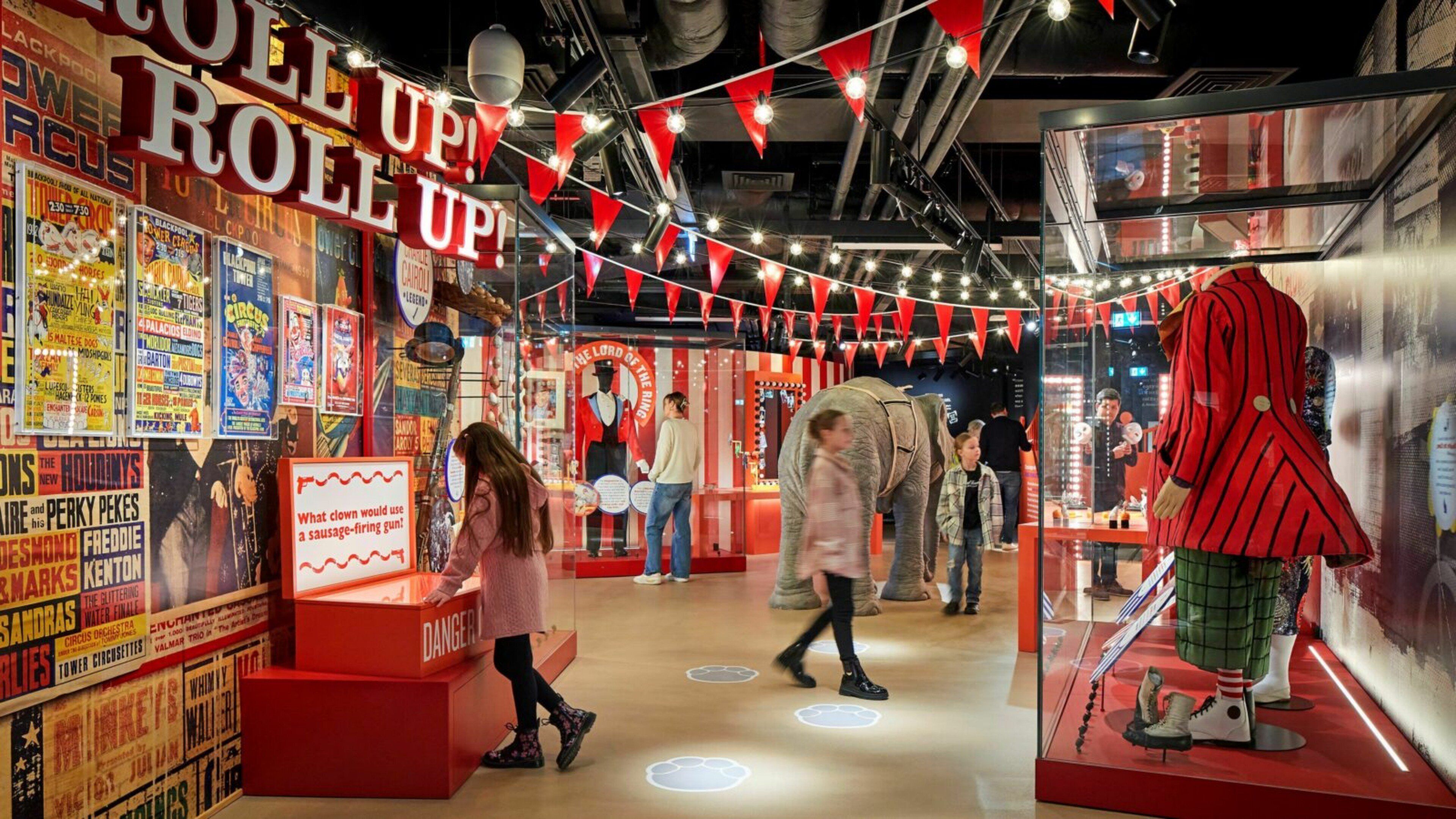 Un espai interior vibrant amb decoració vermella i blanca, que inclou cartells antics, un gran rètol que diu "ROLL UP!", i diverses exposicions, com ara un maniquí amb un vestit vermell i una estàtua d'elefant.