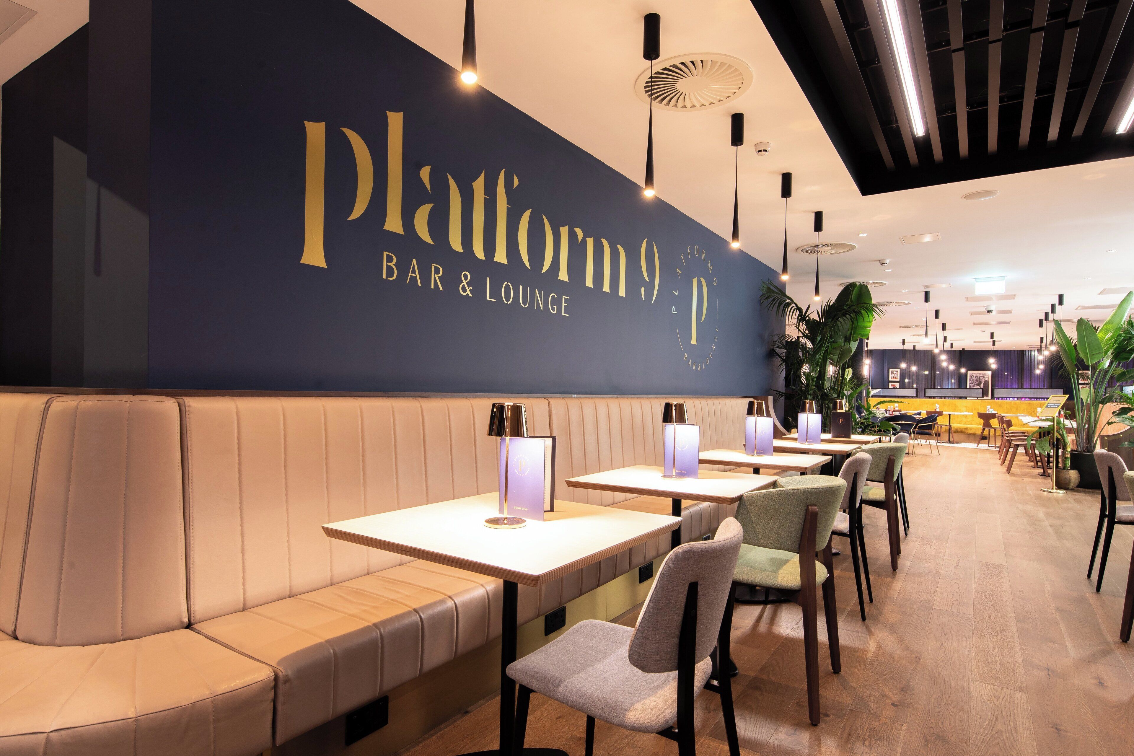 A modern bar and lounge area with a dark wall featuring the name 'Platform 9 BAR & LOUNGE'. There are several tables and chairs arranged for seating.