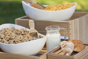Two white bowls filled with granola and cereal, a glass pitcher of milk, and a croissant on a wooden tray.