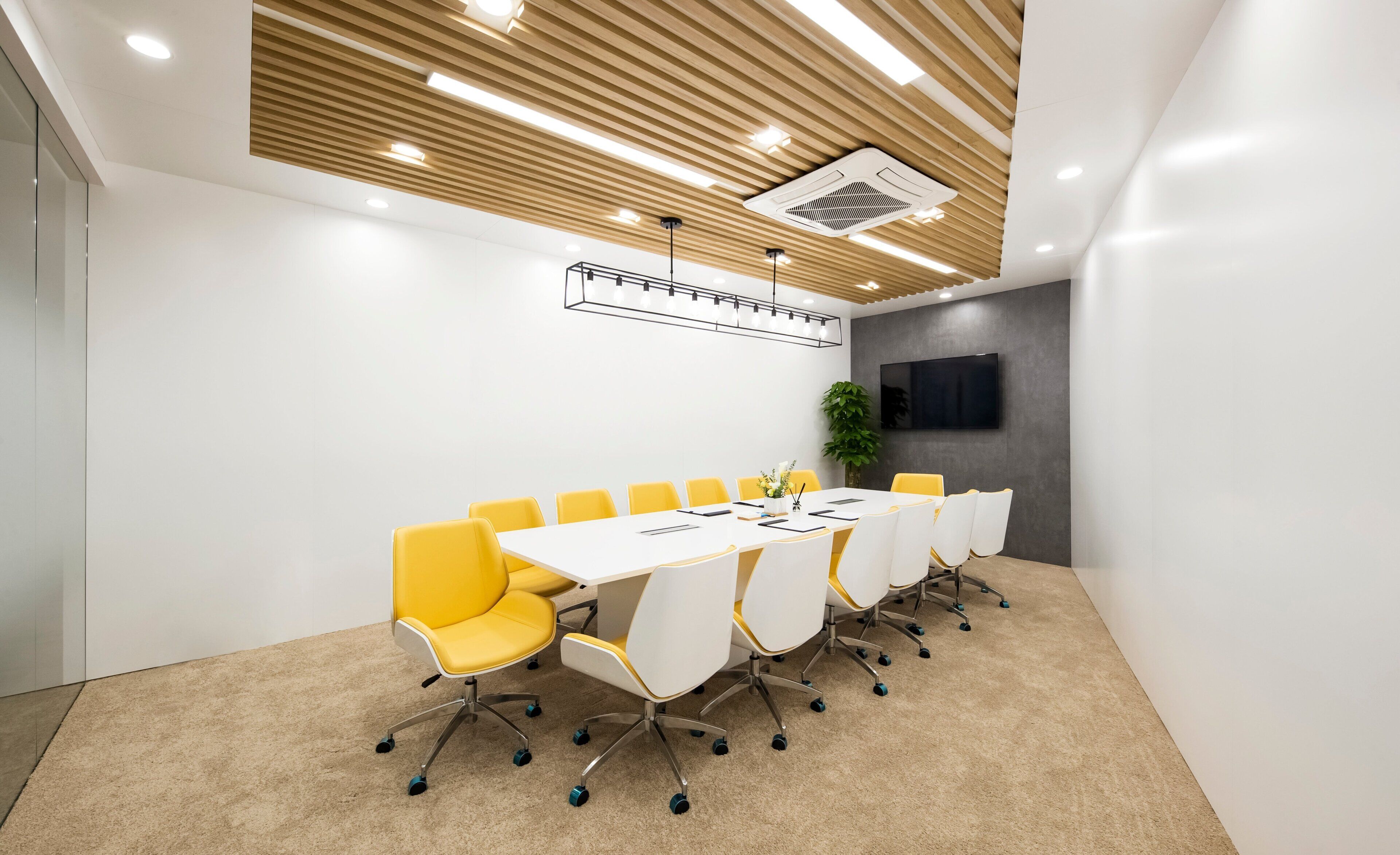 A conference room with a long table, yellow chairs, white chairs, a mounted TV, and a ceiling vent.