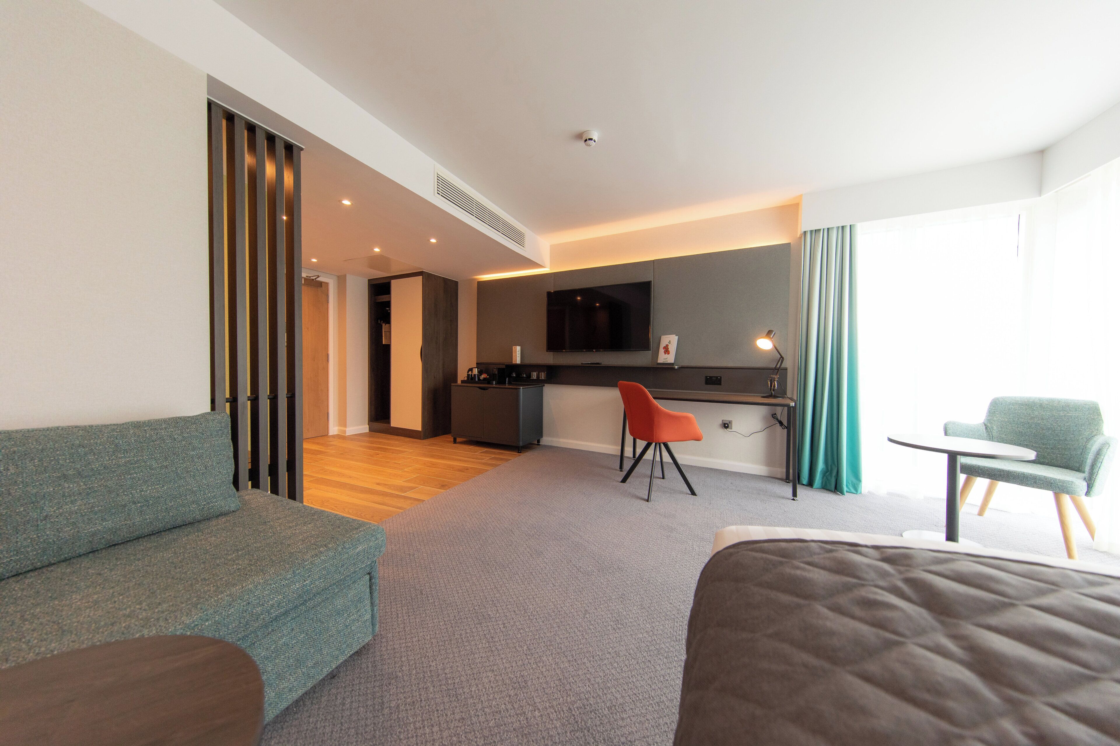A modern hotel room with a sofa, a small dining table, a chair, a TV, and a kitchenette.