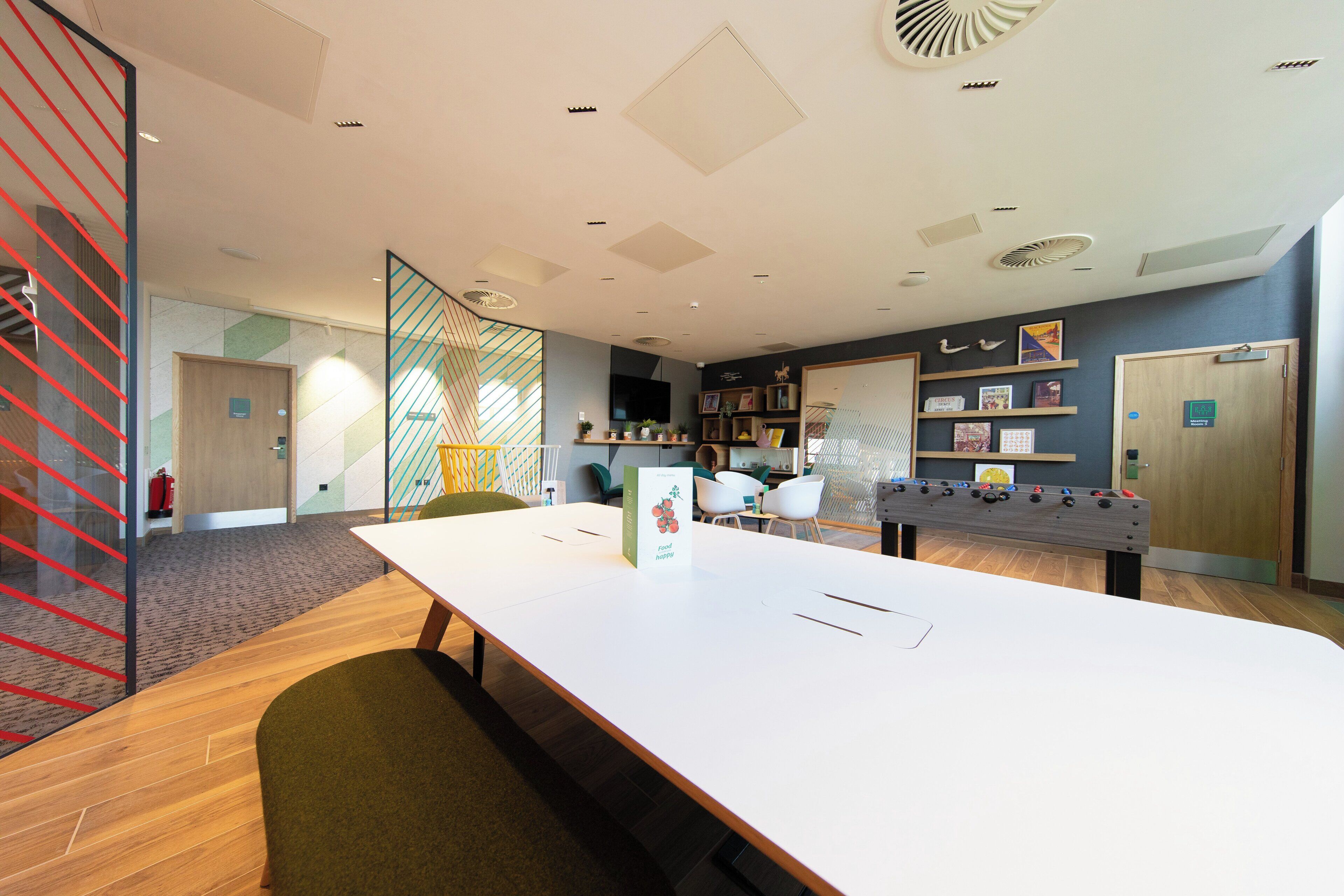 A modern conference room with a long table, chairs, a foosball table, and a bookshelf with various items.