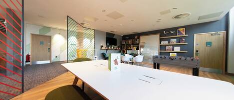 A modern conference room with a long table, chairs, a foosball table, and a bookshelf with various items.