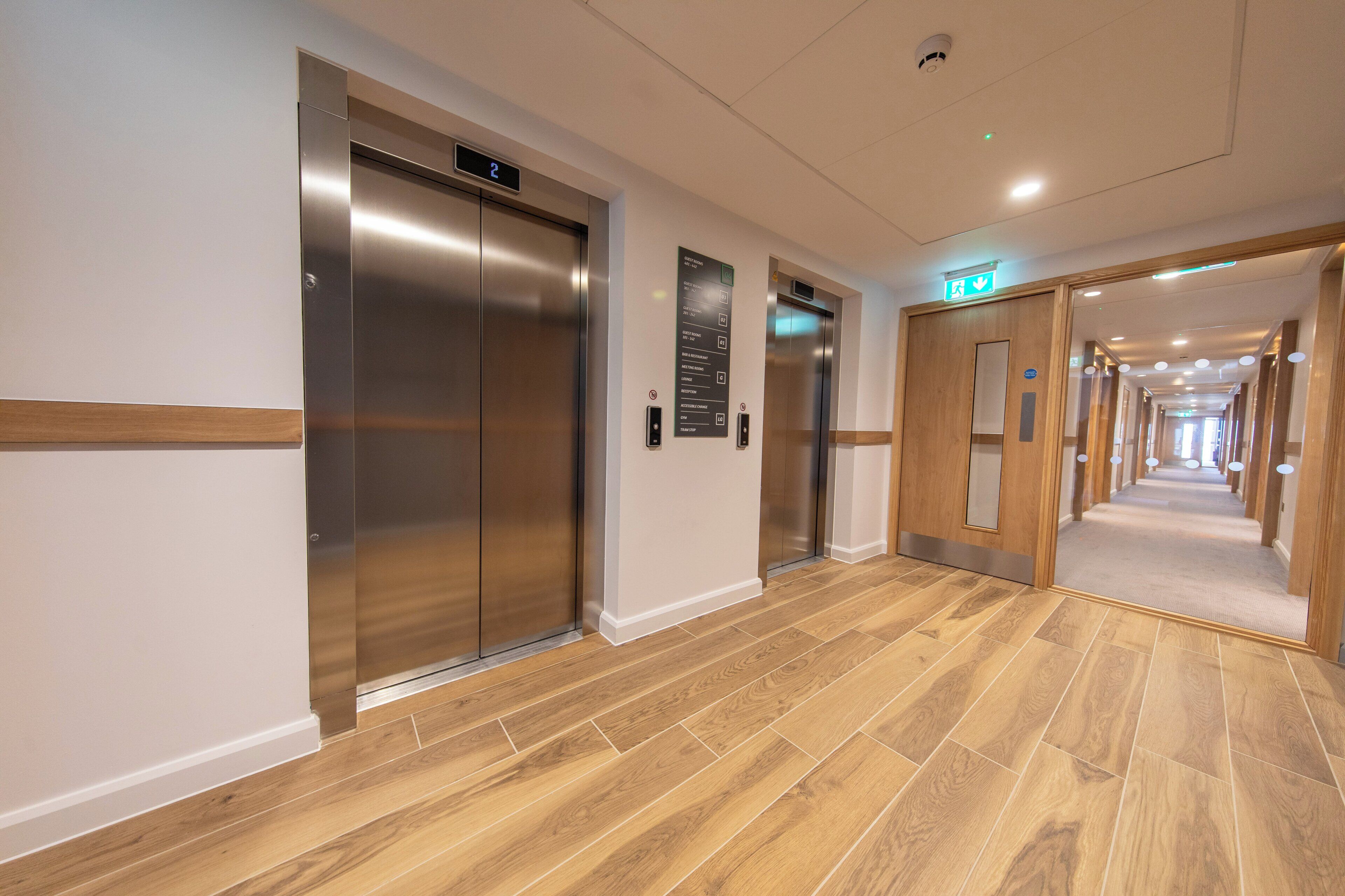 A hotel corridor with two elevators, a wooden floor, and a sign indicating guest rooms.
