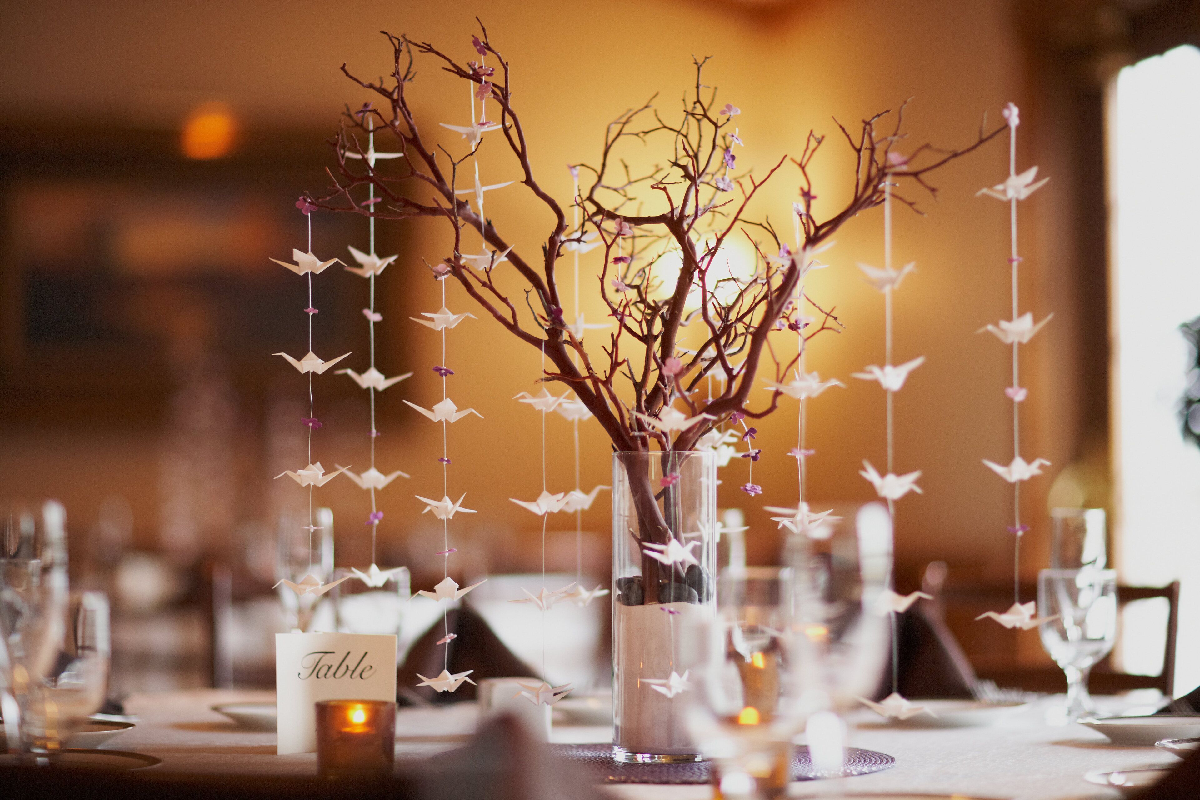 A table set with a vase containing branches and hanging paper birds, a candle, and a table name card.
